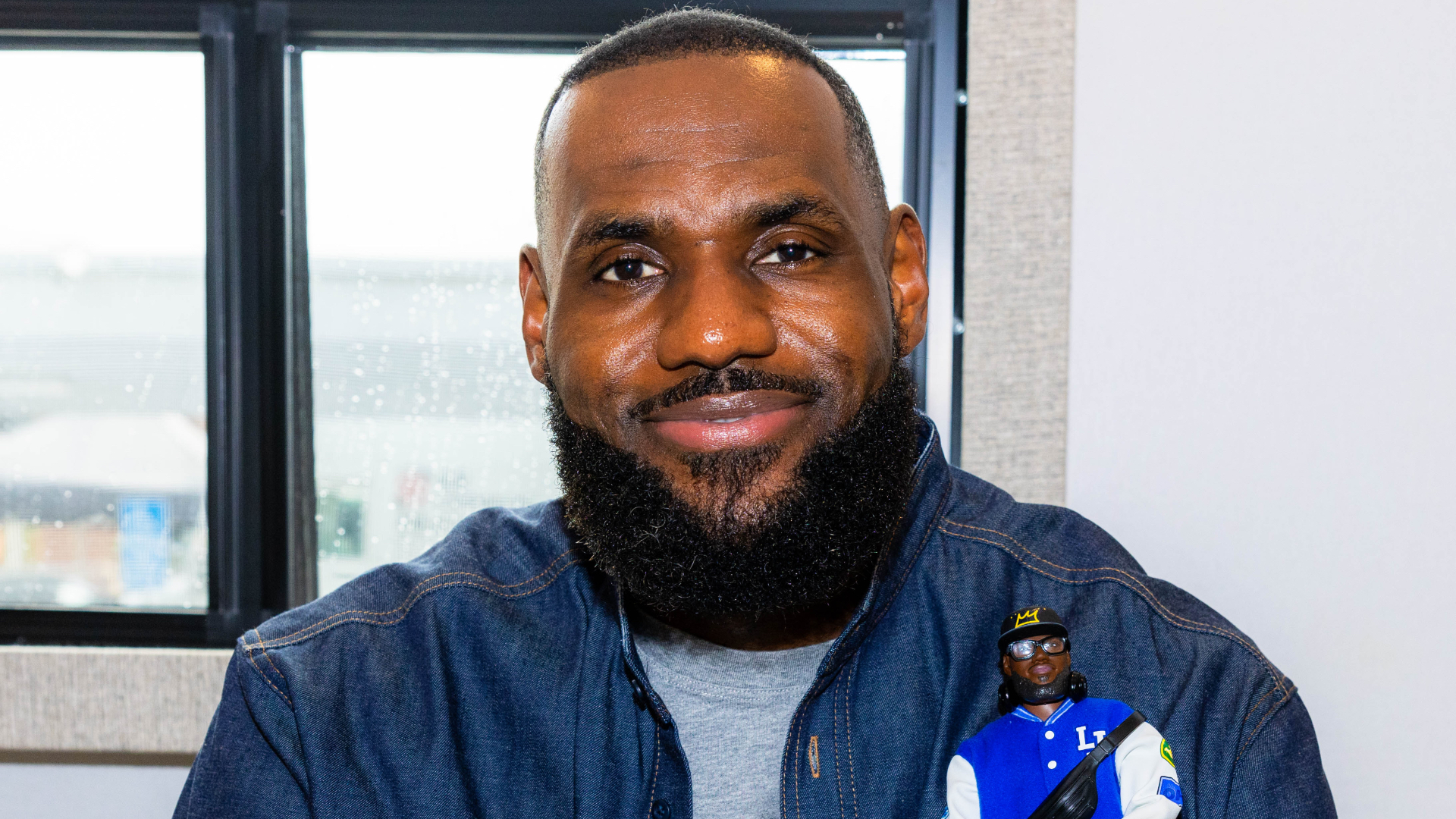 LeBron James smiling, holding a small action figure of himself, wearing a denim jacket, sitting indoors by a window.