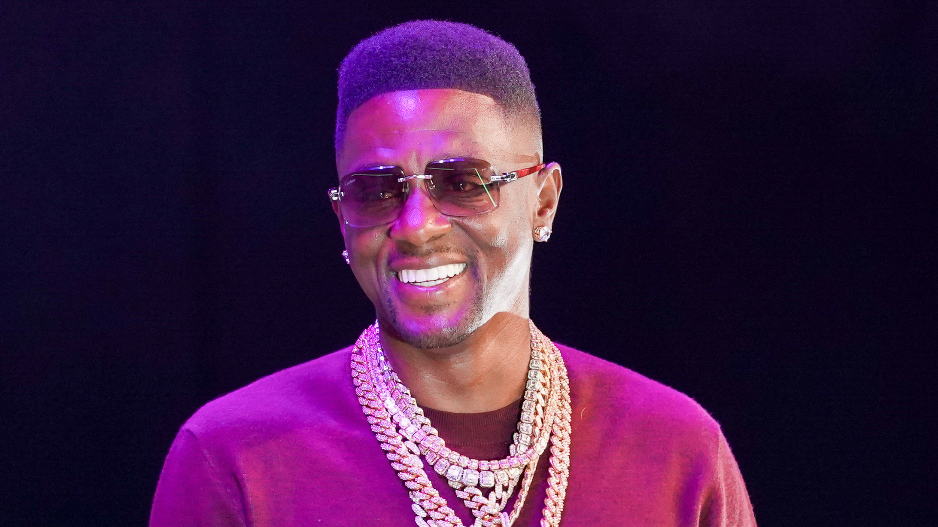 A man wearing sunglasses and multiple gold chains, smiling against a dark background.