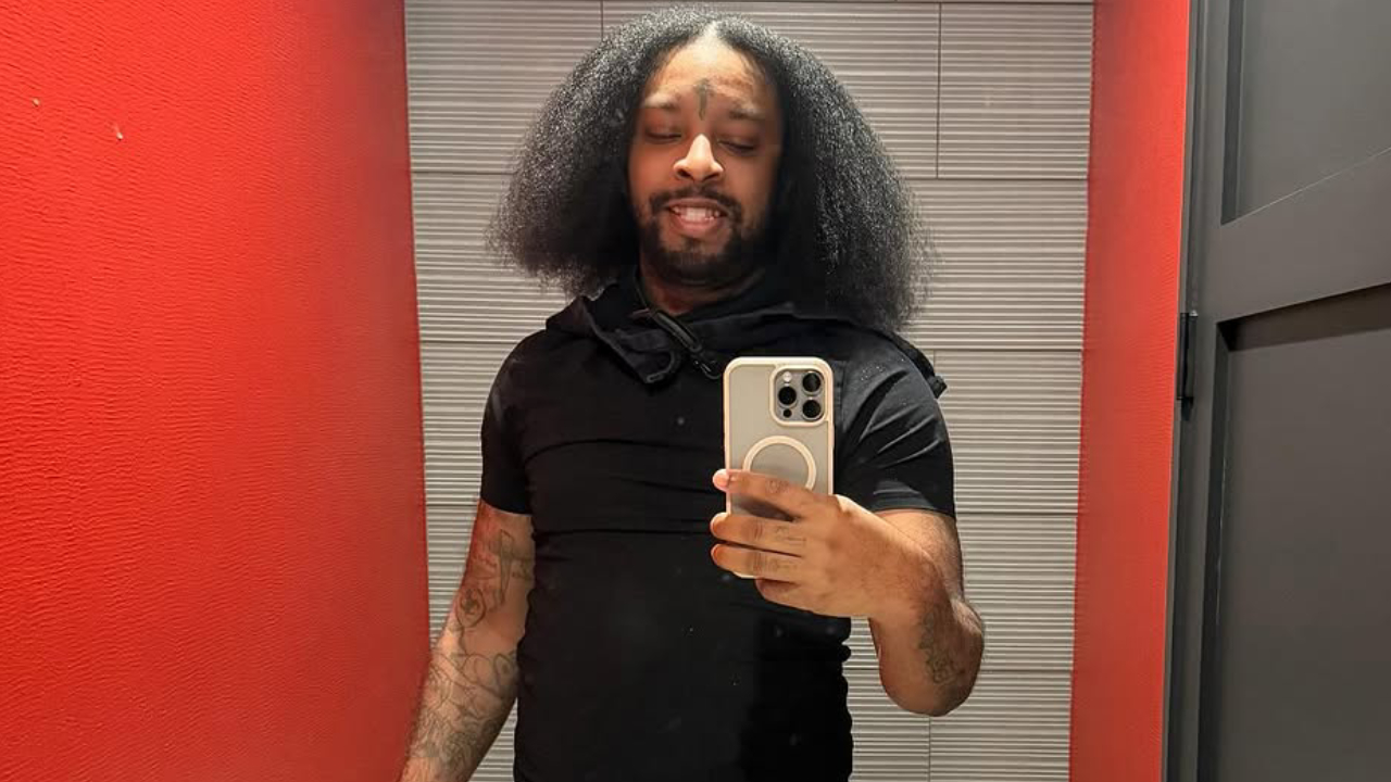 21 Savage with long, curly hair takes a mirror selfie in a room with red walls, wearing a black shirt and holding a smartphone.