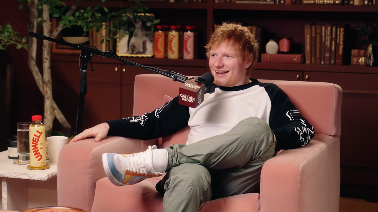 Ed Sheeran sitting in a pink chair, smiling, with a microphone. He's wearing a black and white shirt and green pants.