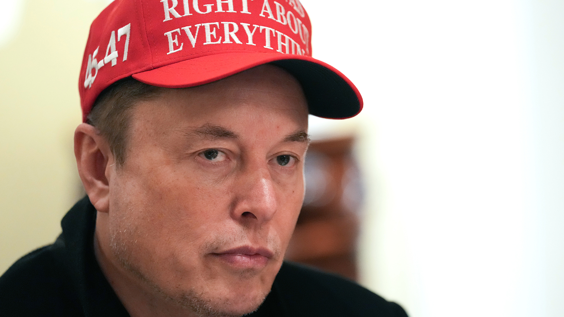 Elon Musk wearing a red MAGA cap with white text, looking mindless, against a blurred background.