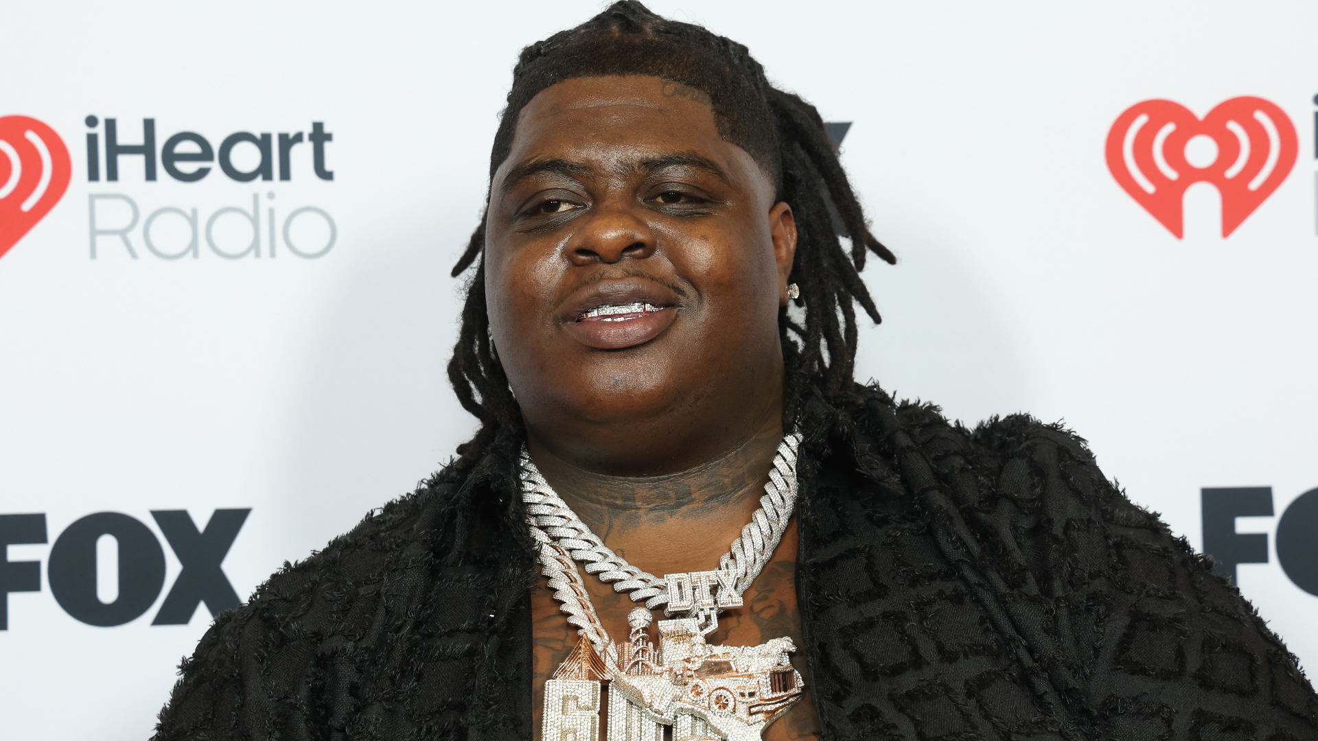 BigXthaPlug with dreadlocks and a black jacket, wearing large silver necklaces, stands in front of an iHeartRadio and FOX backdrop.