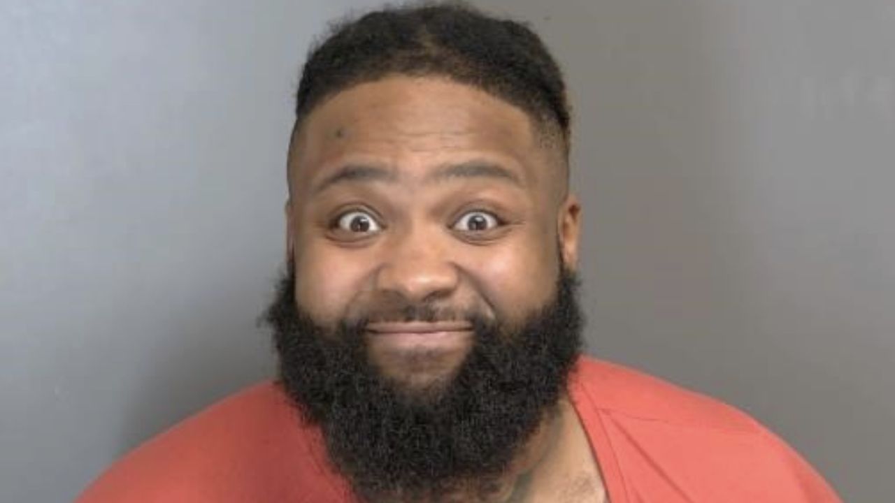 Sada Baby with a beard and wide eyes wearing an orange shirt against a plain gray background.