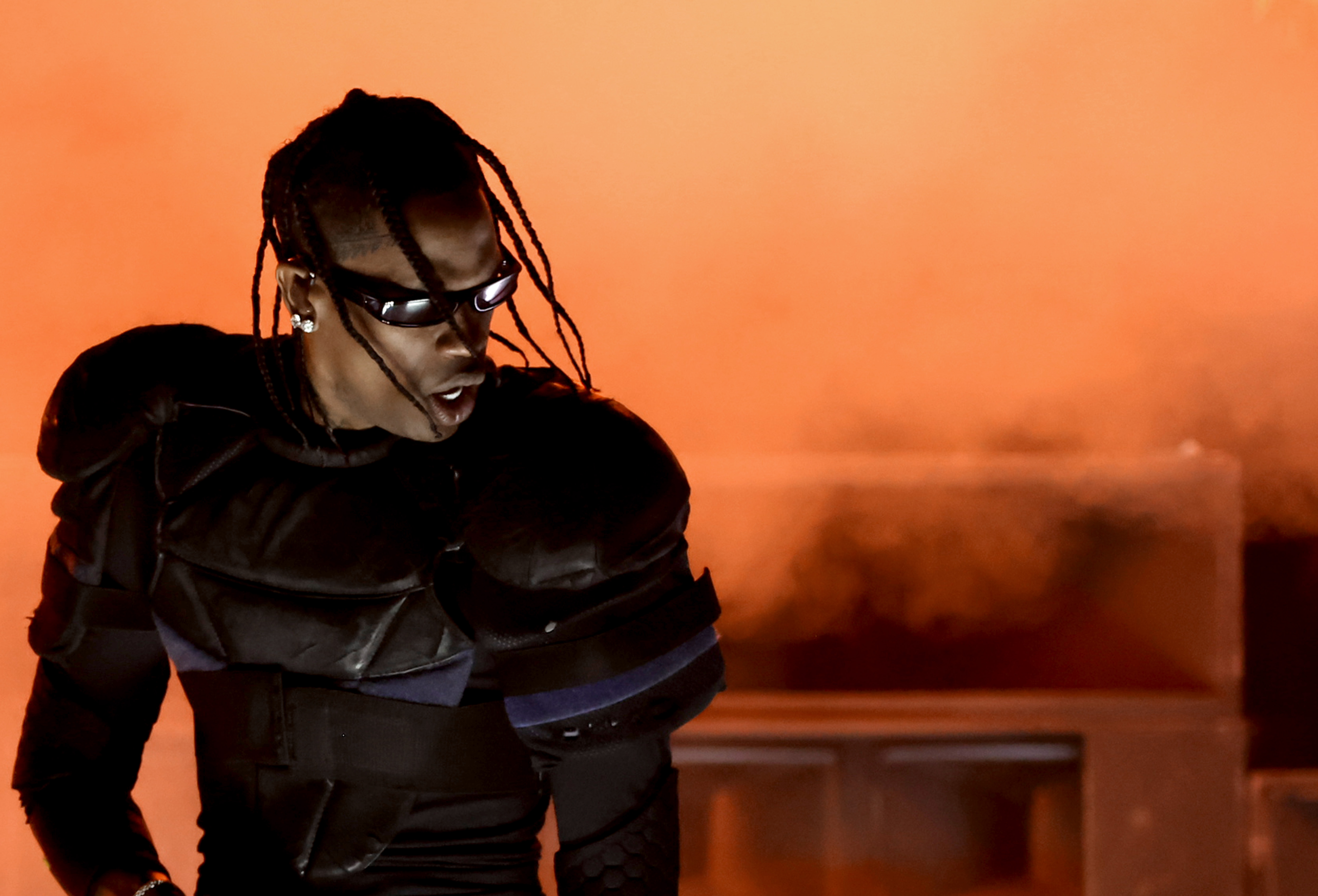 A performer in futuristic black armor and sunglasses stands in front of an orange smoky background.