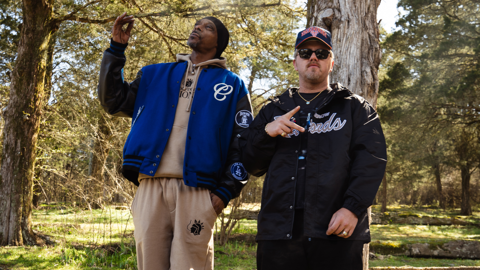 Snoop Dogg and Ernest standing outdoors, Snoop in a blue jacket and beige pants, Ernest in a black jacket and cap, with trees behind them.