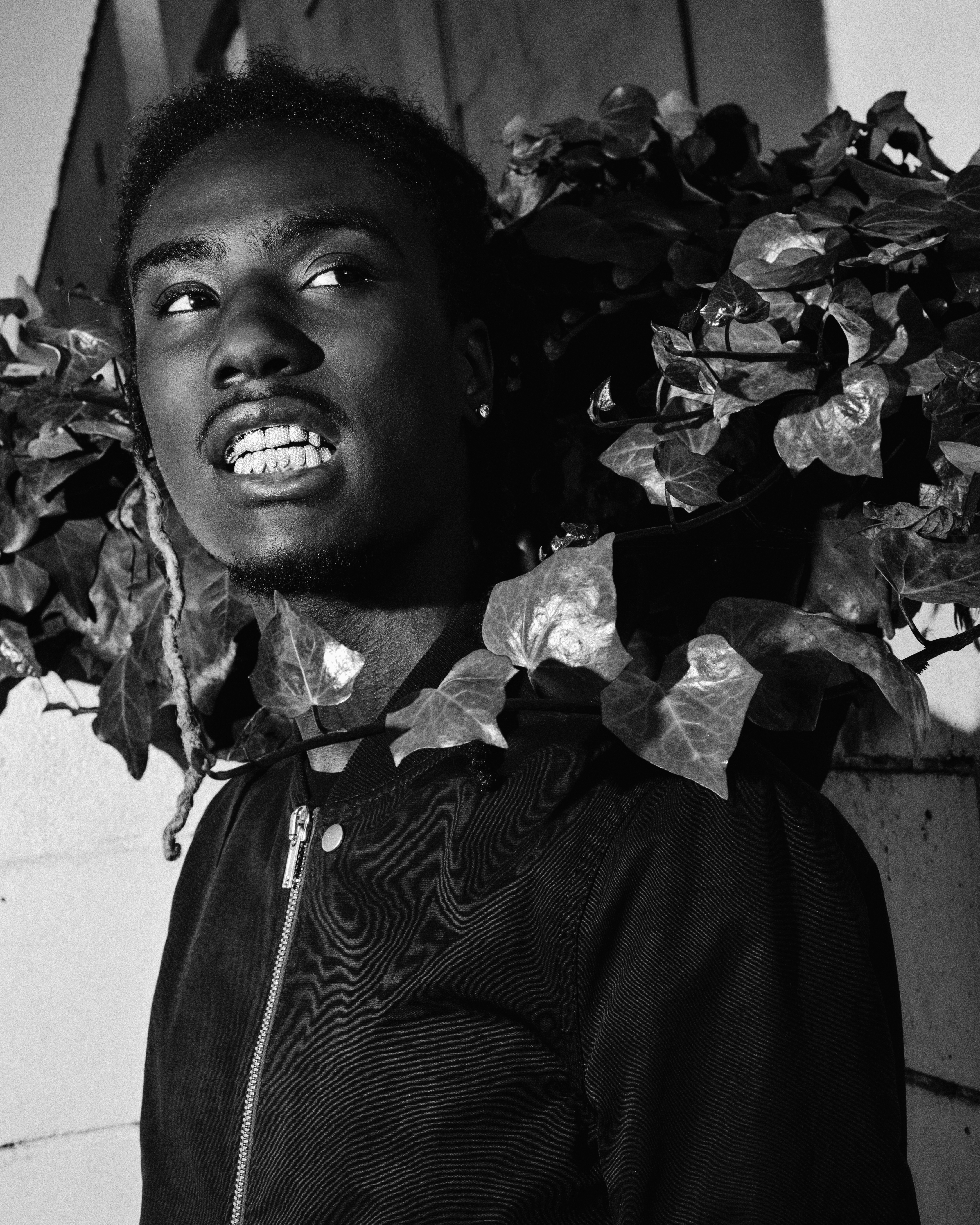 A black and white image of OsamaSon with grills, wearing a jacket, surrounded by leaves.