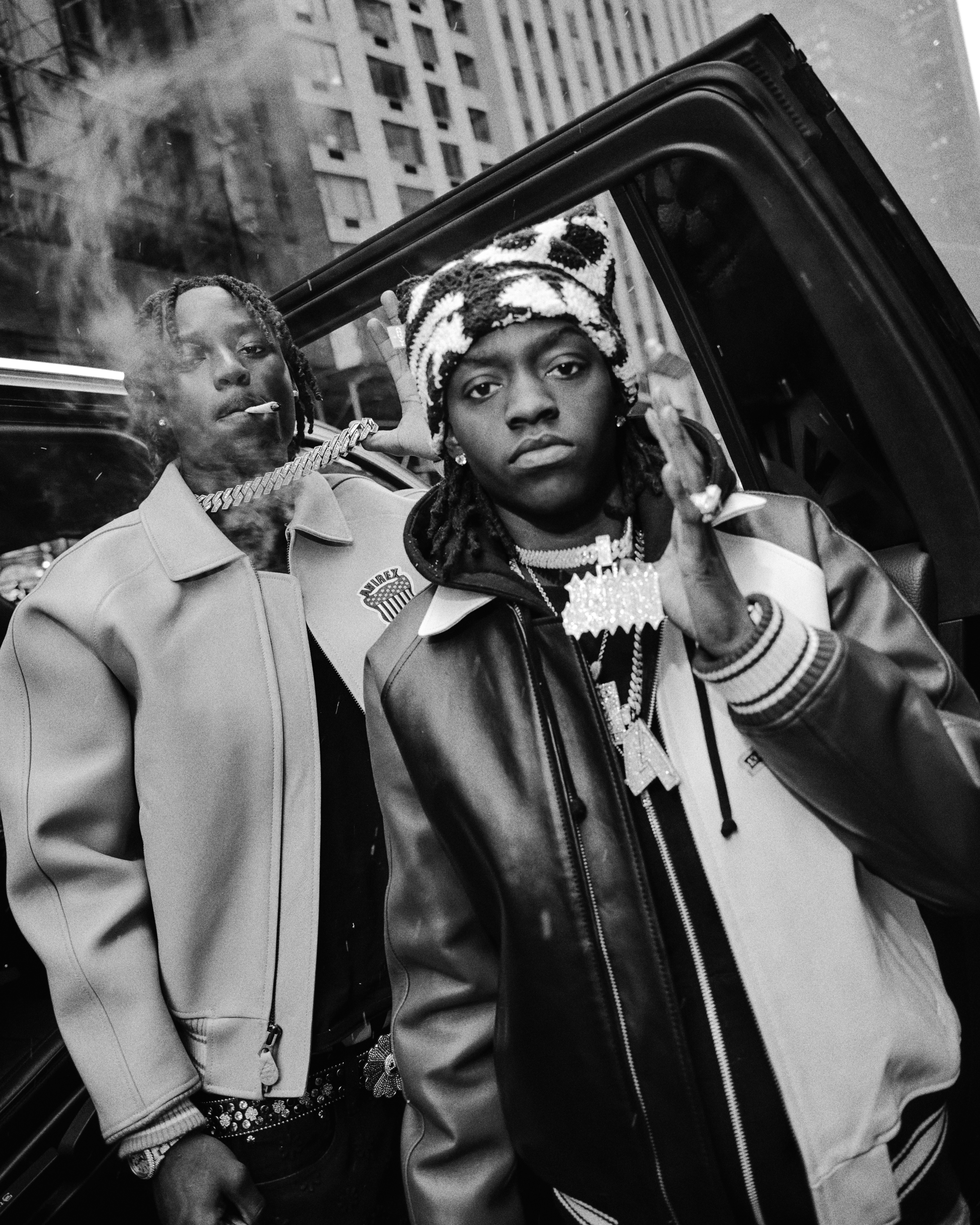 41 in stylish jackets stand by a car in a city. One is smoking, and both wear jewelry. Black and white photo.