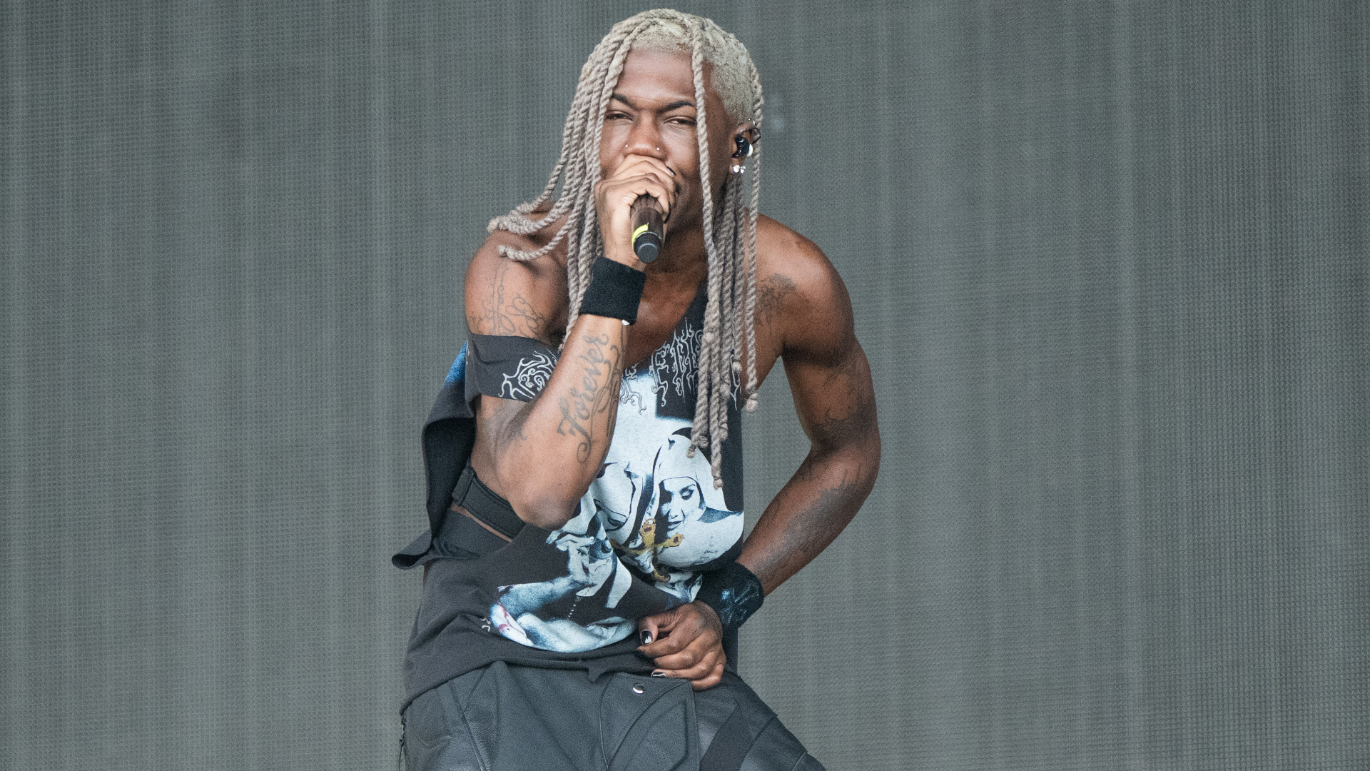 Destroy Lonely with long, braided hair and tattoos sings passionately on stage, wearing a graphic tank top and black pants.