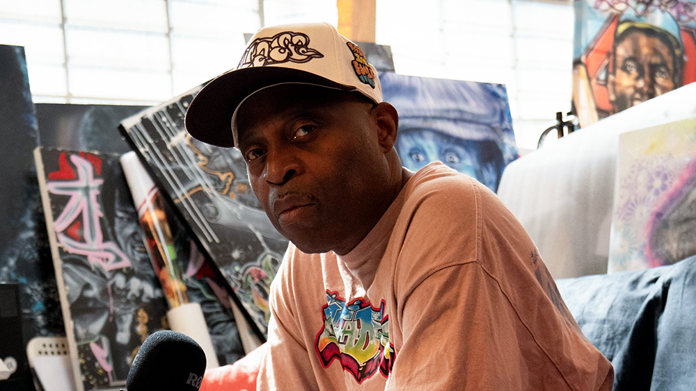 Shirt King Phade in a cap and graphic t-shirt sits in a room filled with colorful artwork, looking intently at the camera.