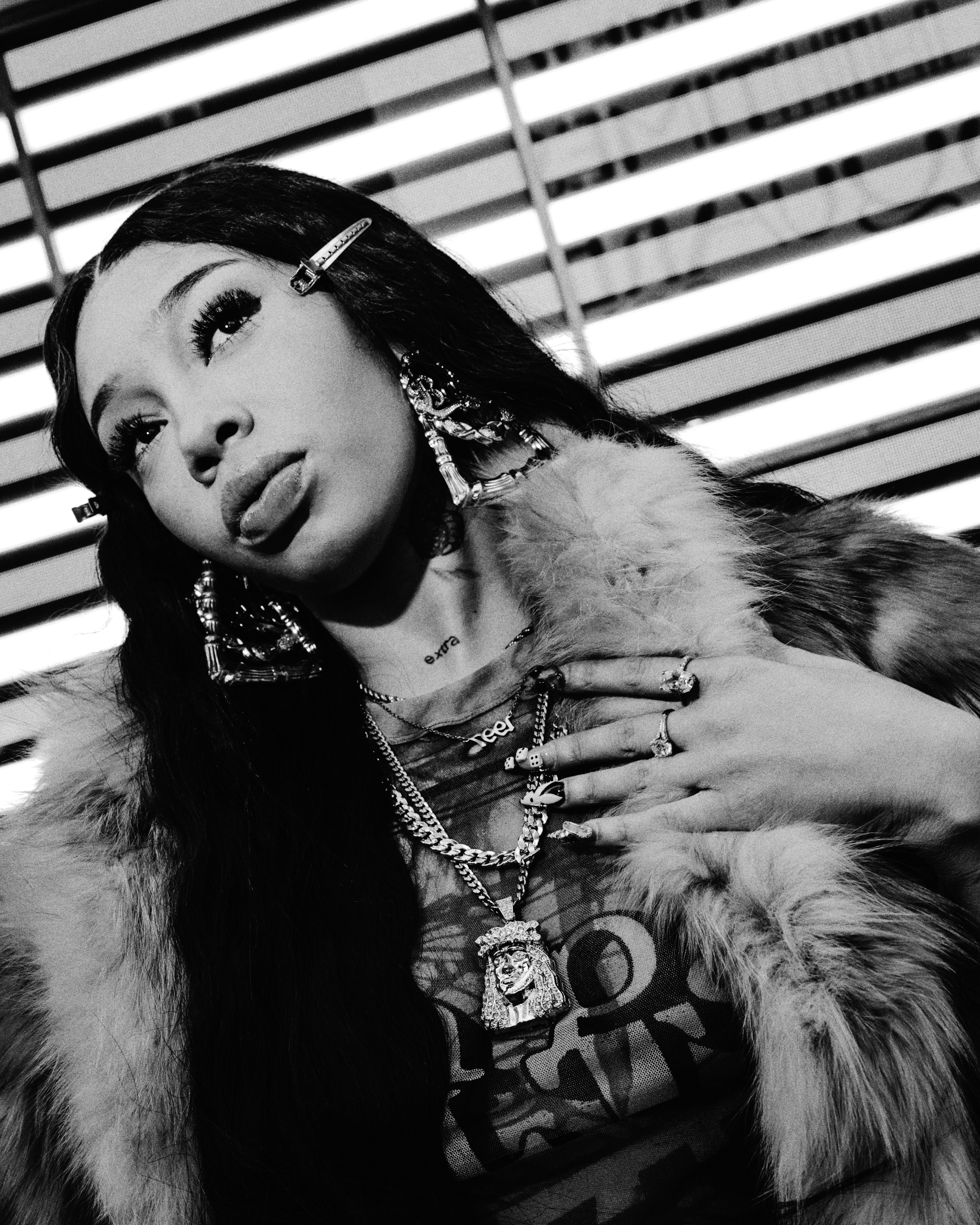 A woman with long hair, wearing fur, large earrings, and layered necklaces, poses in front of horizontal blinds. Black and white image.