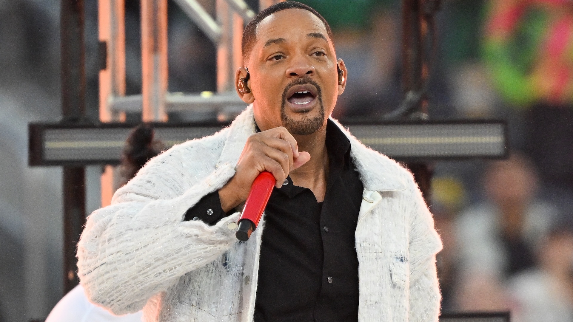 Will Smith performing on stage, wearing a light jacket and holding a red microphone.