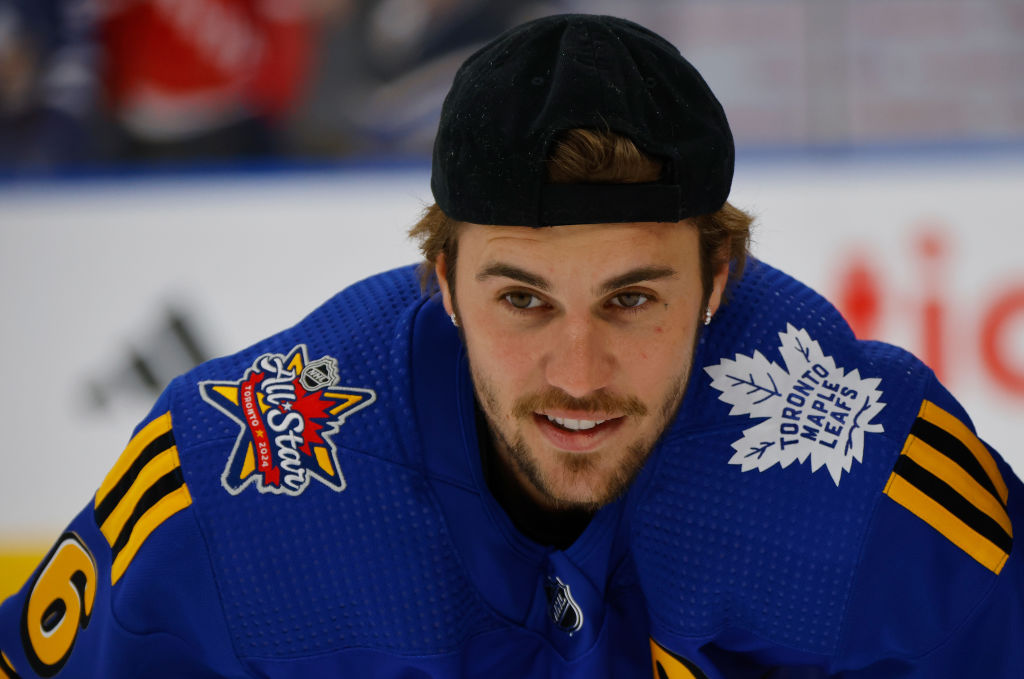 Justin Bieber in a blue jersey with Toronto Maple Leafs logo and All-Star patch, wearing a black cap backward, smiling.