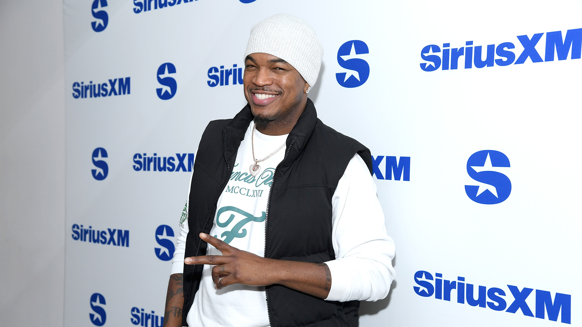 Ne-Yo at a SiriusXM event, wearing a white beanie, black vest, and white shirt, smiling and making a peace sign.