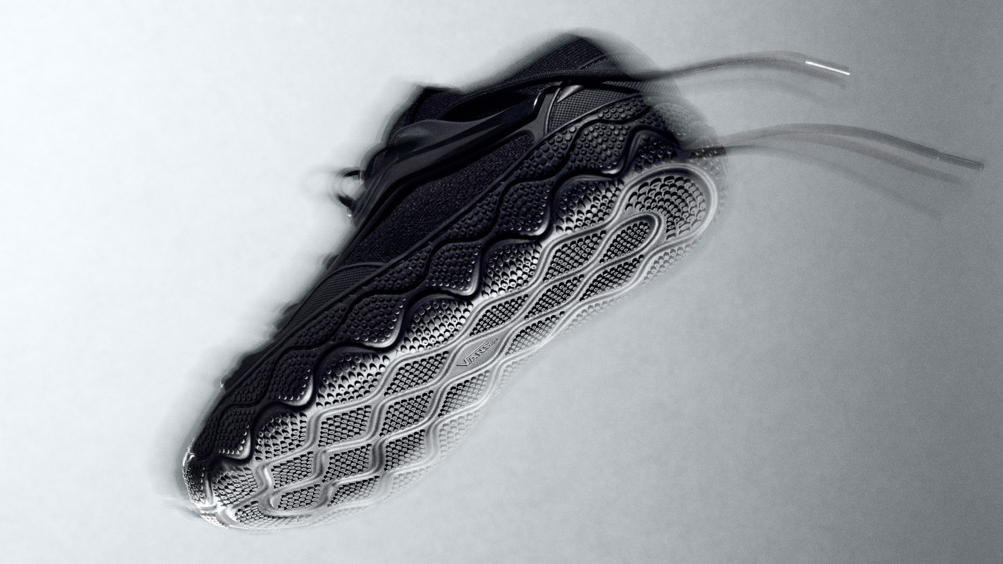 A black sneaker with a textured, wavy sole design, photographed from below on a gray background.
