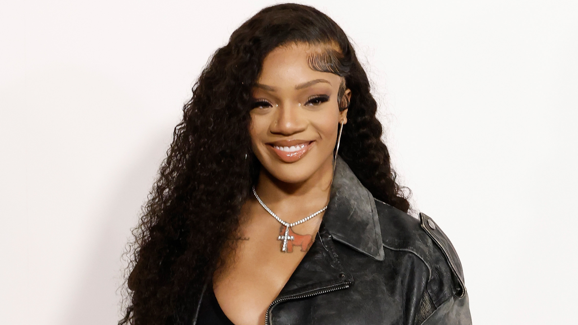 A woman with long curly hair, wearing a black leather jacket and a cross necklace, smiles against a plain background.
