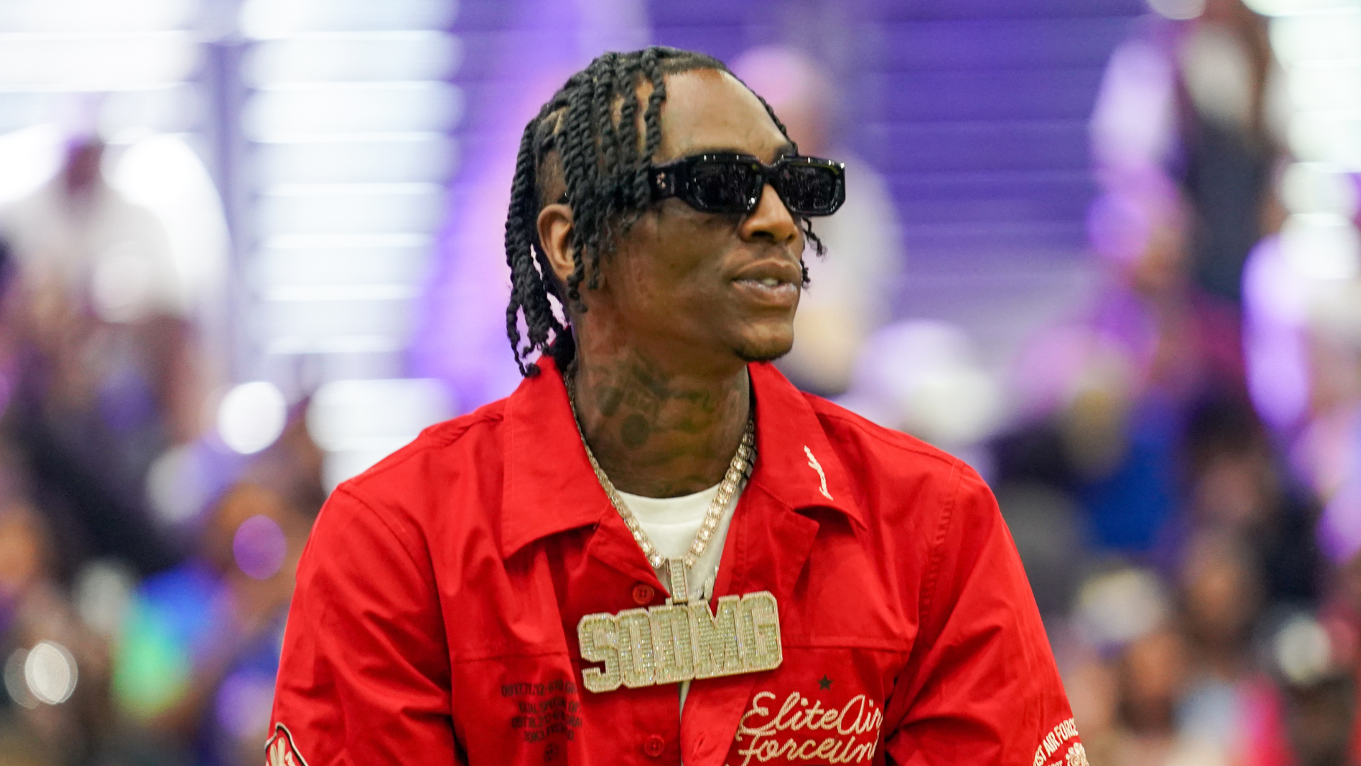 Soulja Boy wearing a red shirt, sunglasses, and a large chain necklace, with braided hair, in a lively indoor setting.