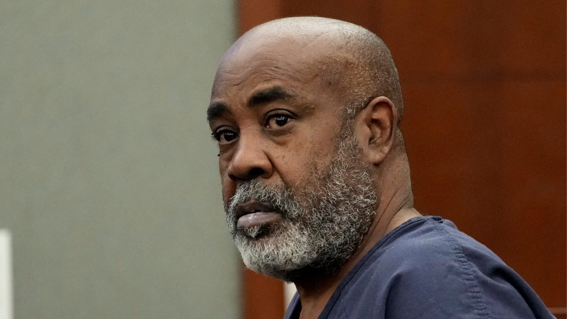 A man with a gray beard and wearing a dark shirt looks to the side in a courtroom setting.