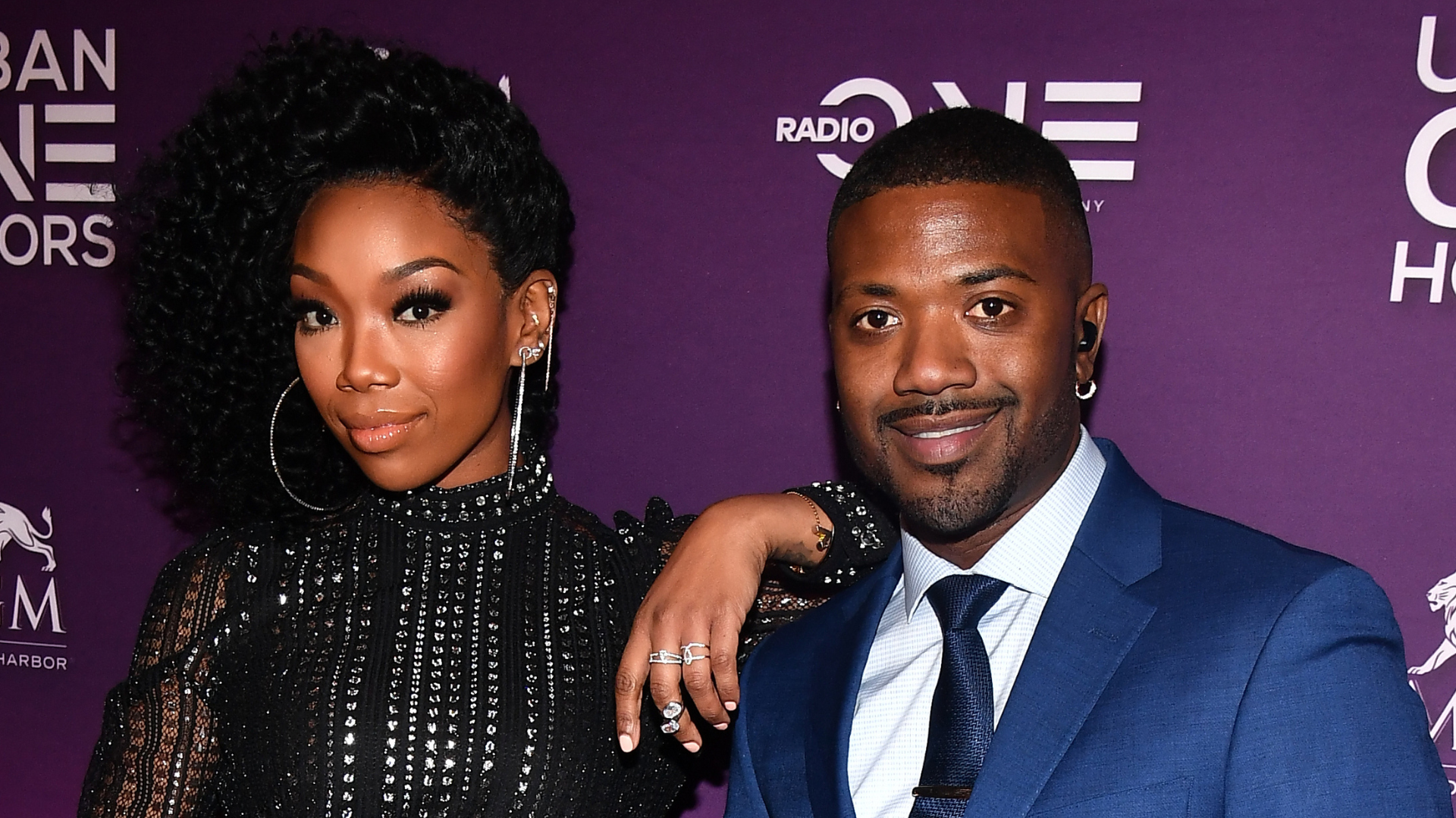 Brandy and Ray J at an event, posing together. Brandy is in a black dress, and Ray J is in a blue suit.