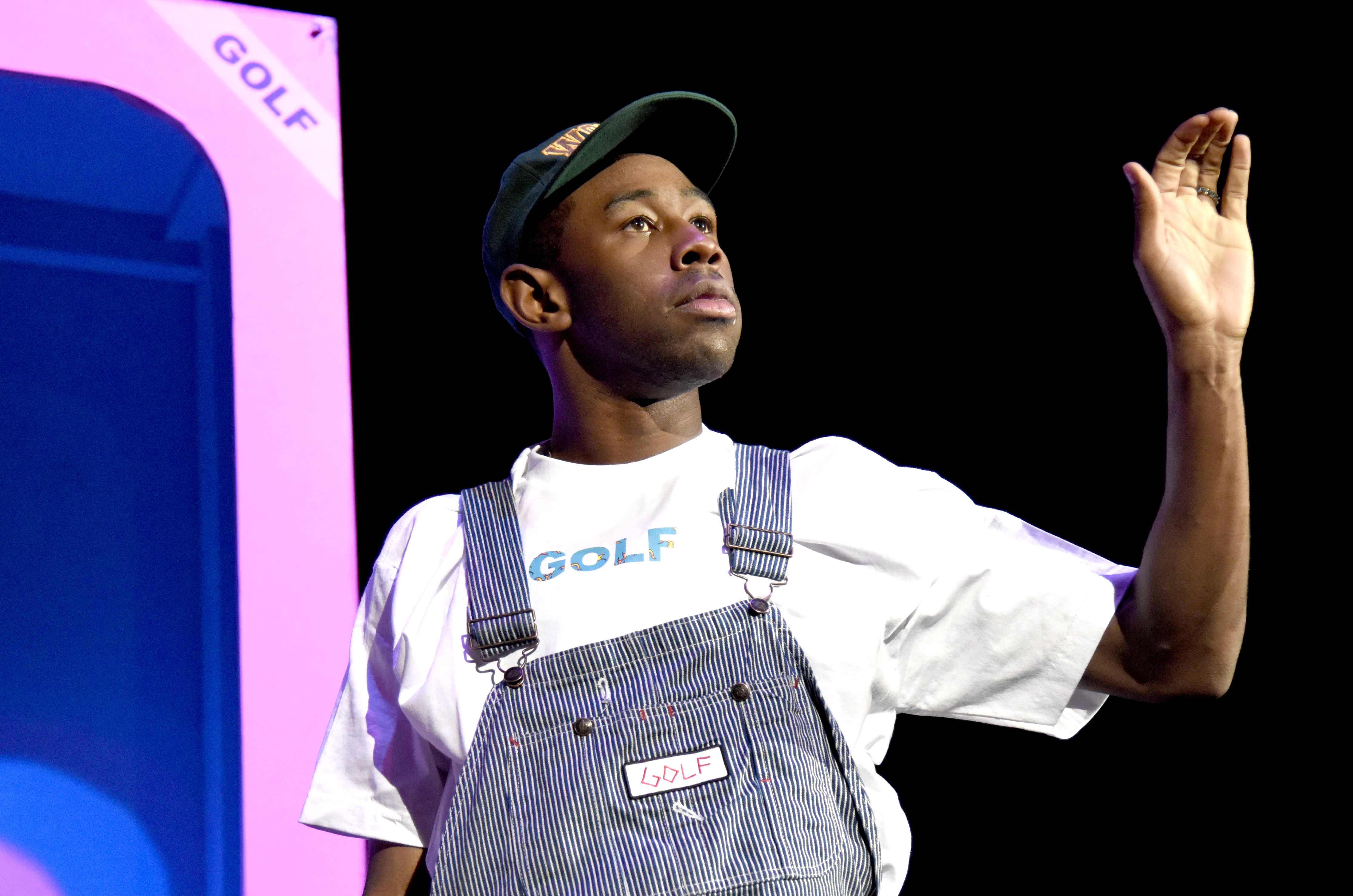 Tyler, the Creator wearing a "GOLF" shirt and overalls, waving on stage with a colorful backdrop.