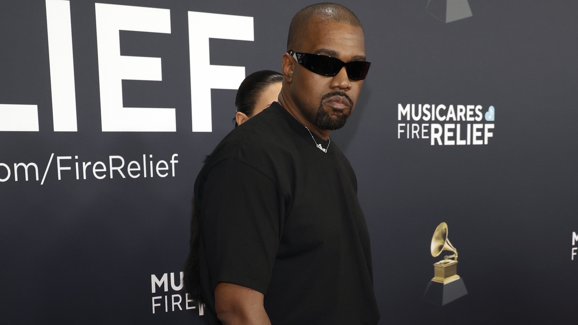 Kanye West wearing sunglasses and a black shirt at a MusiCares event, standing against a backdrop with event branding.