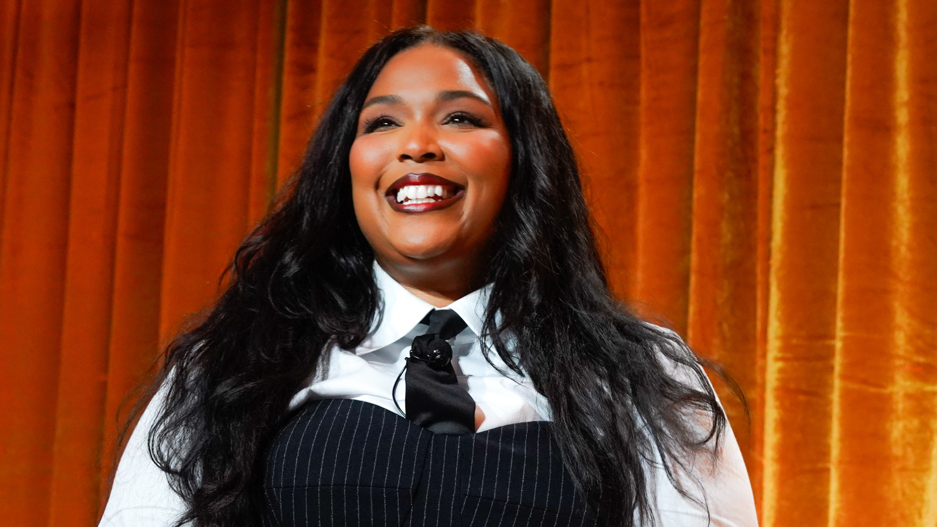 Lizzo smiling in front of an orange curtain, wearing a white shirt and black pinstripe vest with a black tie.
