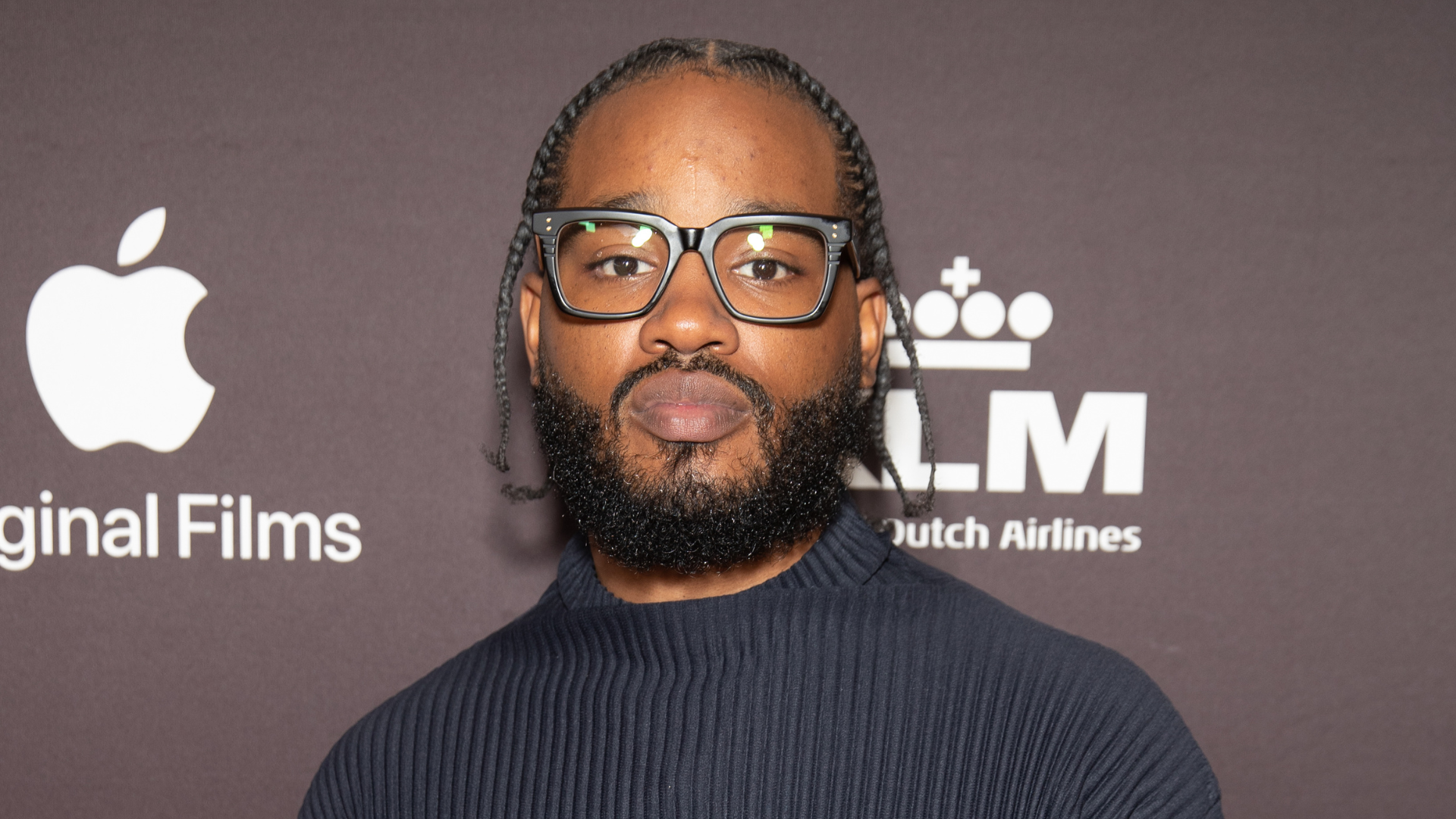 A man with glasses and a beard at an event, with Apple Original Films and KLM logos in the background.