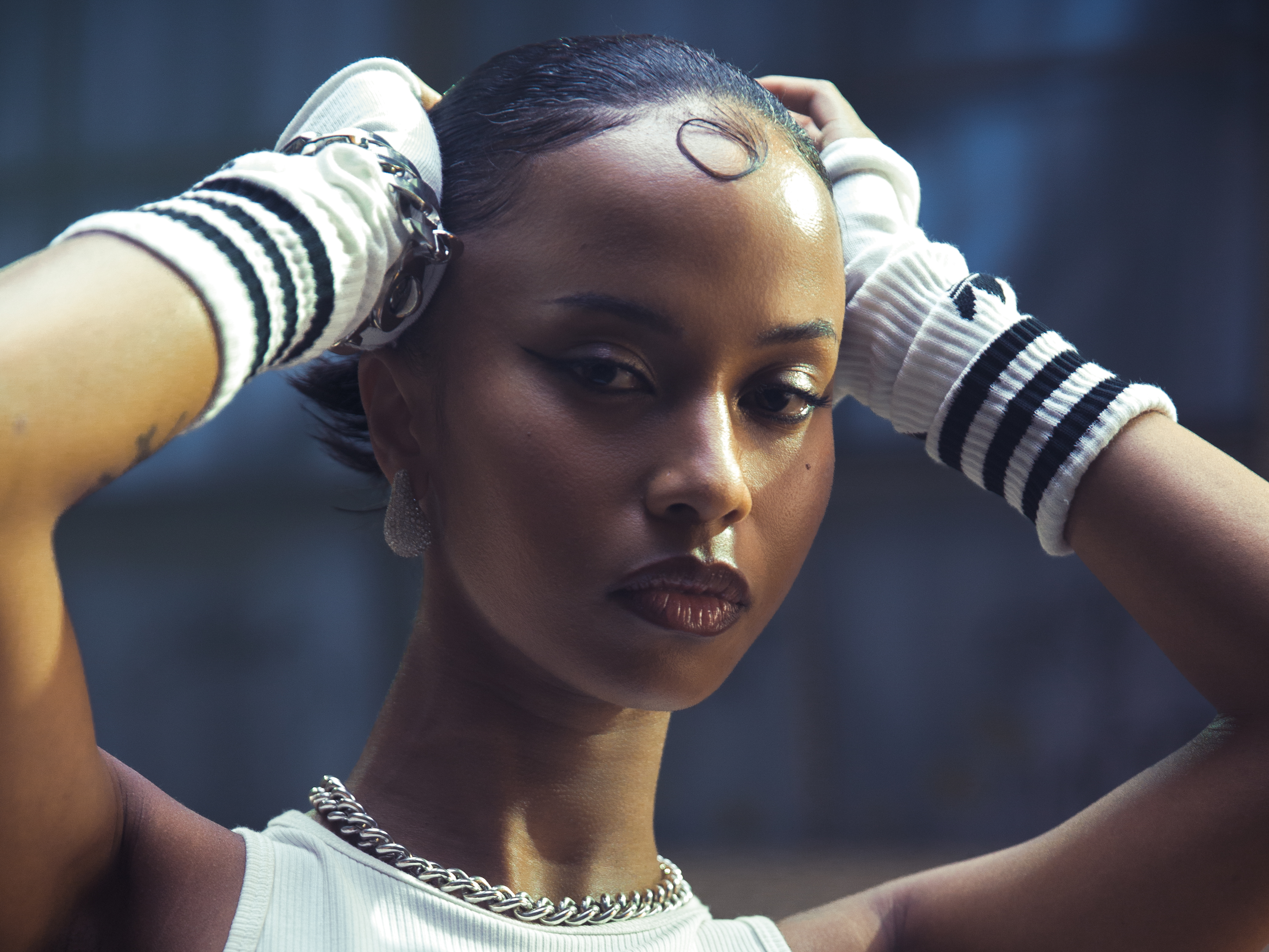 A woman with slicked-back hair, wearing a white top and silver jewelry, gazes intensely at the camera.