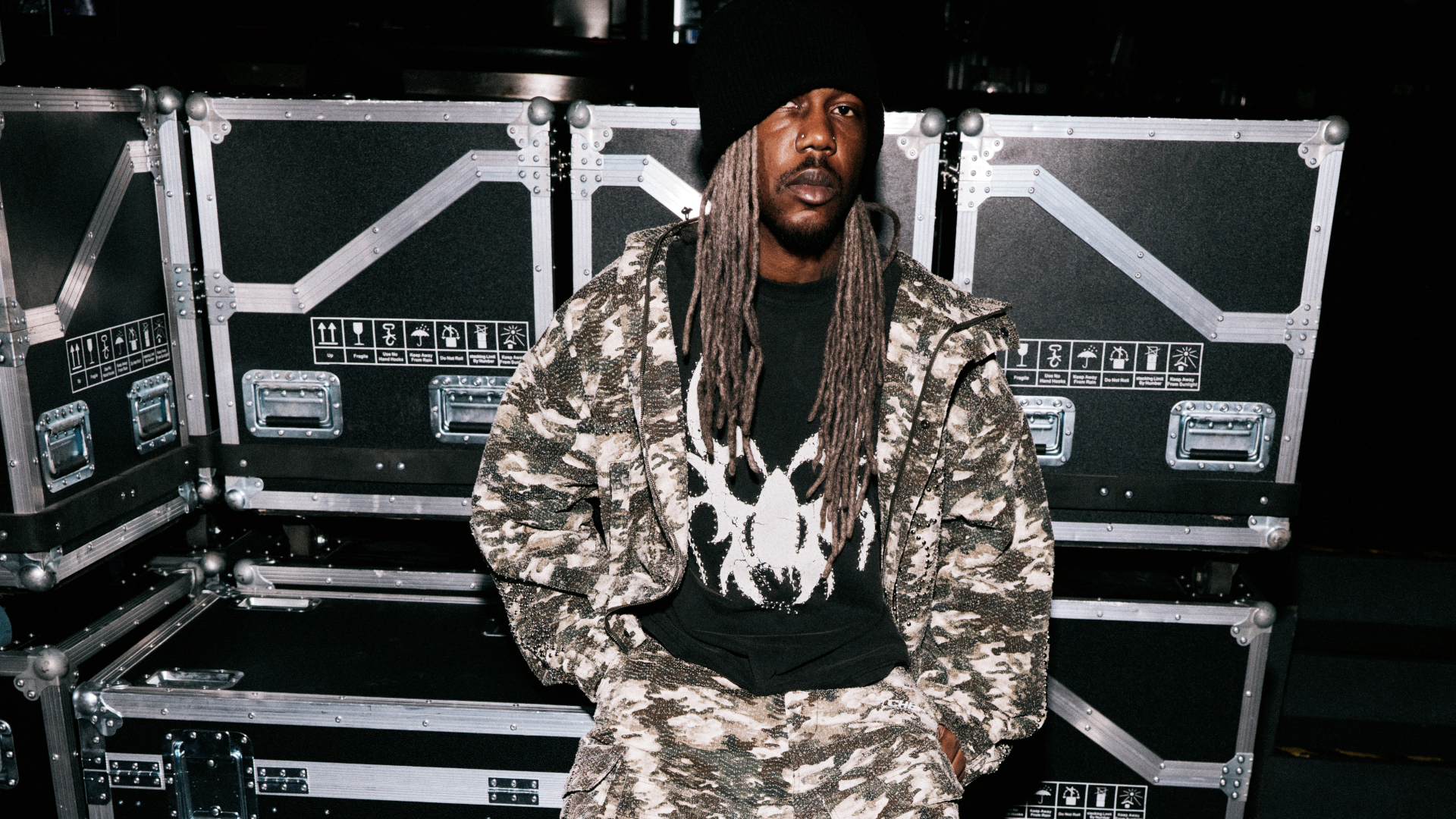 A person with long dreadlocks wearing a patterned outfit and beanie sits in front of stacked equipment cases.
