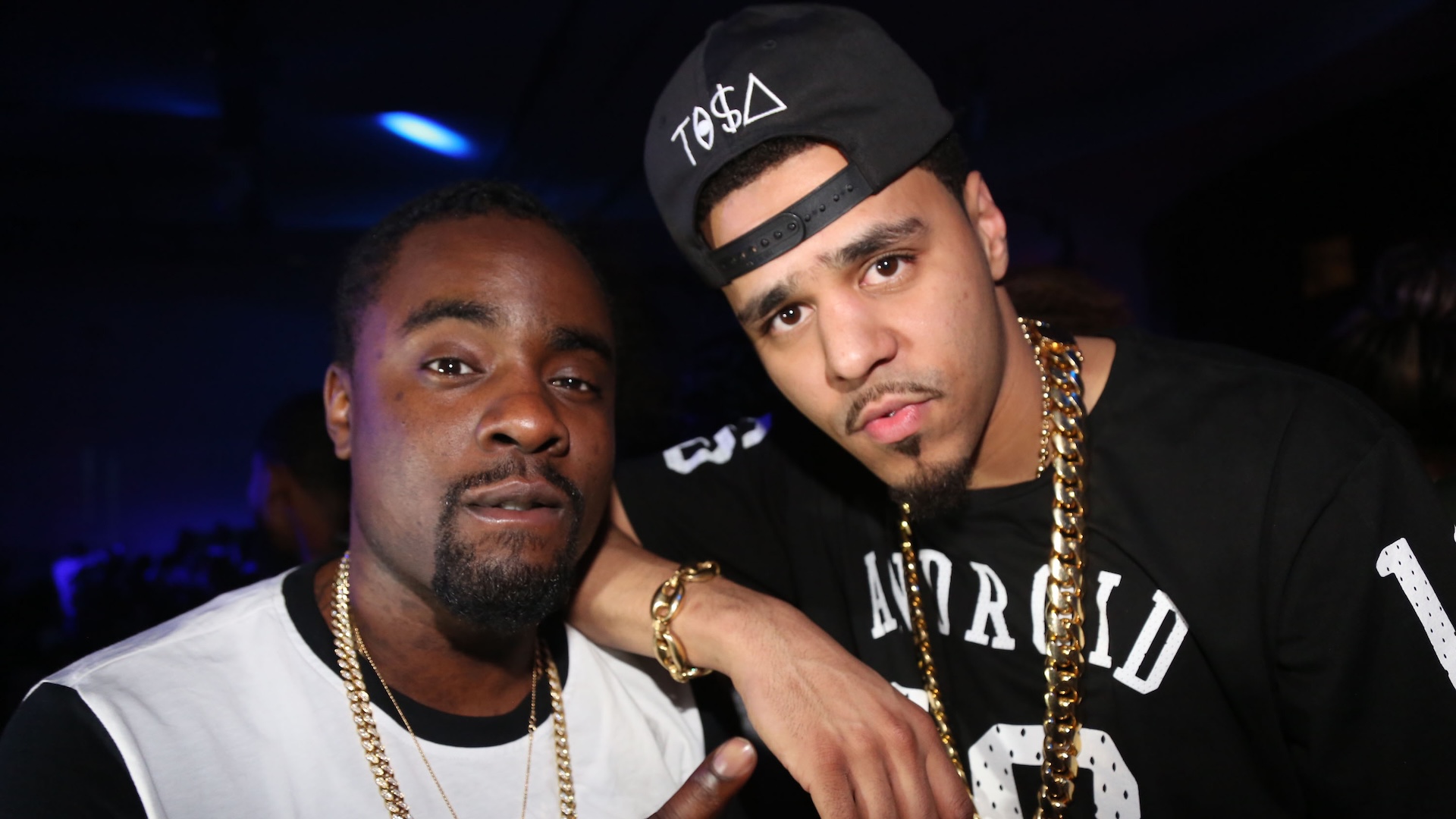 J. Cole is dressed in a black jersey and a black backwards baseball cap. His right hand is placed on Wale's shoulder. Wale is wearing a white shirt with a black neckline and sleeves, pointing at J. Cole. Both artists are staring at the camera.