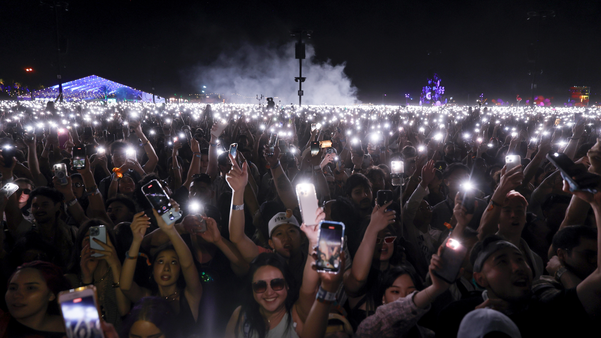 A large crowd at a concert, holding up phones with flashlights on, creating a sea of lights against the night sky.