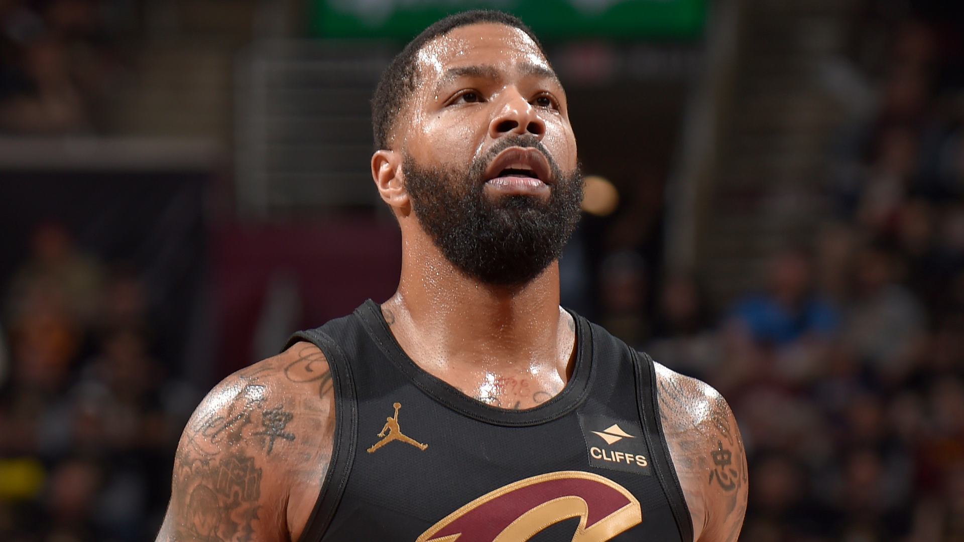 Basketball player in a black Cleveland Cavaliers jersey on the court, with tattoos on his arms, looking focused.