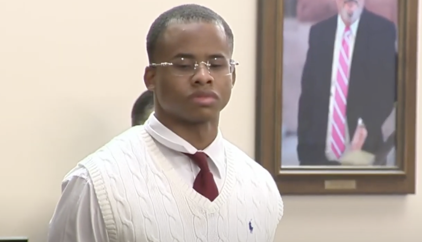 A man in a white sweater and glasses stands in a courtroom, with a framed photo of a man in a suit behind him.