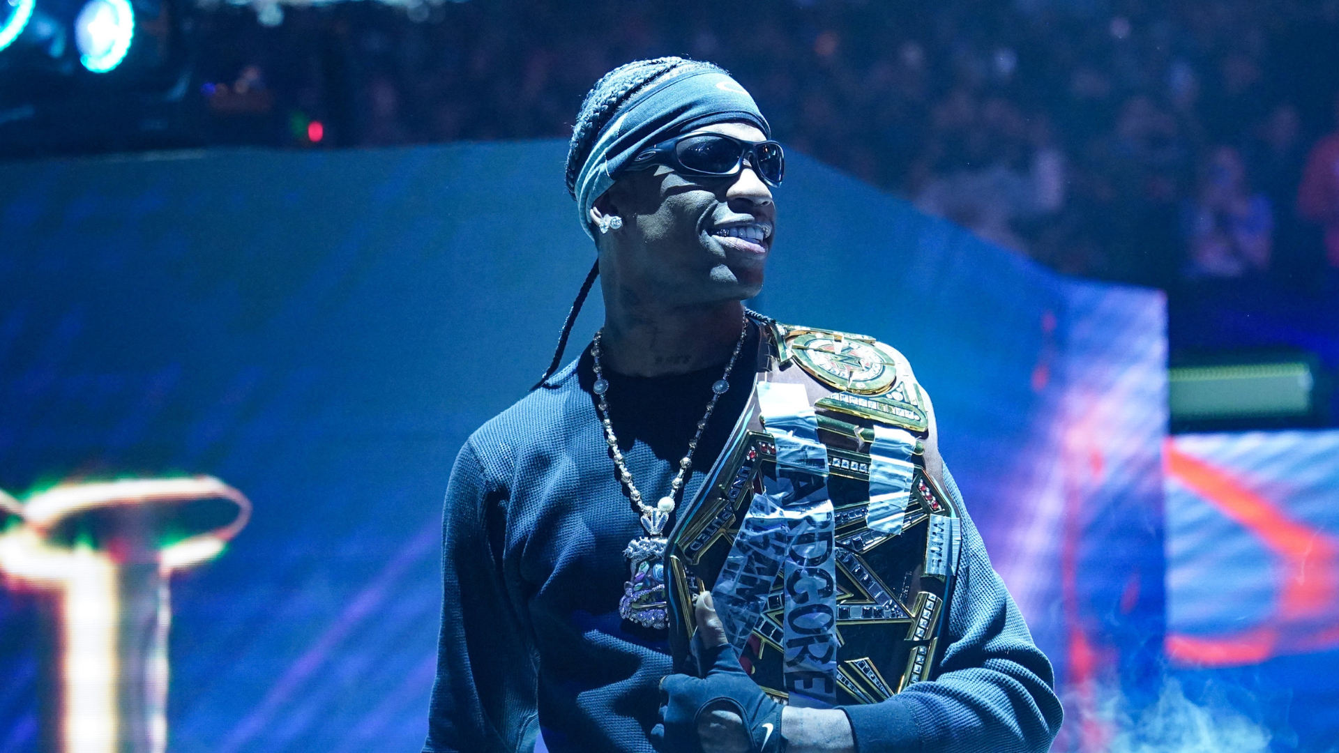 A man wearing sunglasses and a headband, holding a championship belt, stands confidently in a dramatic, smoke-filled setting.