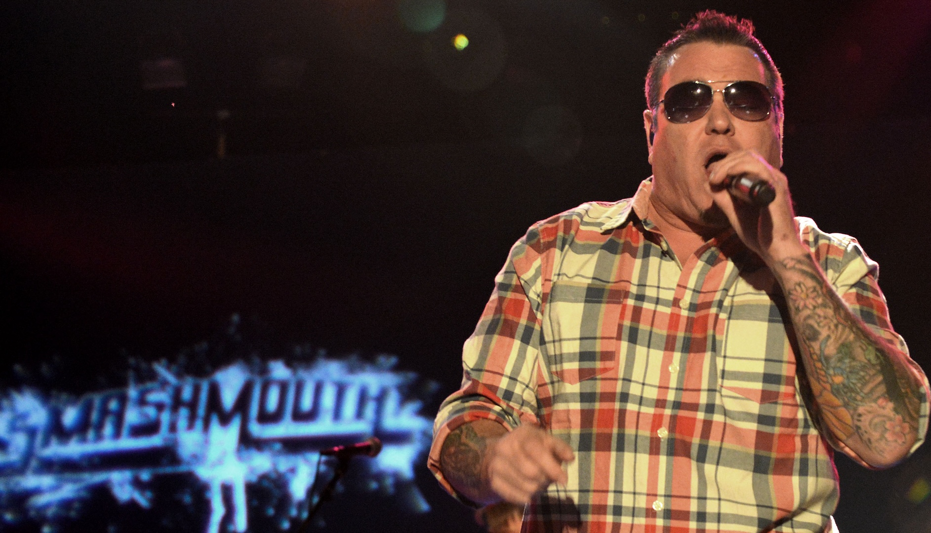 A man in sunglasses and a plaid shirt sings on stage with a microphone. The background displays the word "Smash Mouth."