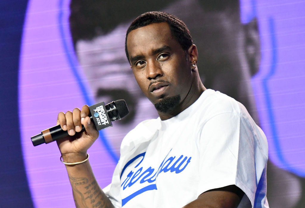 Sean "Diddy" Combs holding a microphone, wearing a white "Crenshaw" shirt, on stage with a serious expression.
