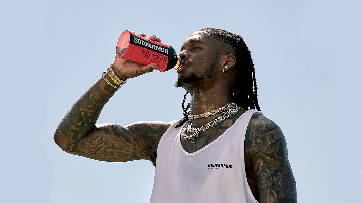 CeeDee Lamb drinks from a BodyArmor bottle, wearing a white tank top and necklaces, against a clear sky.