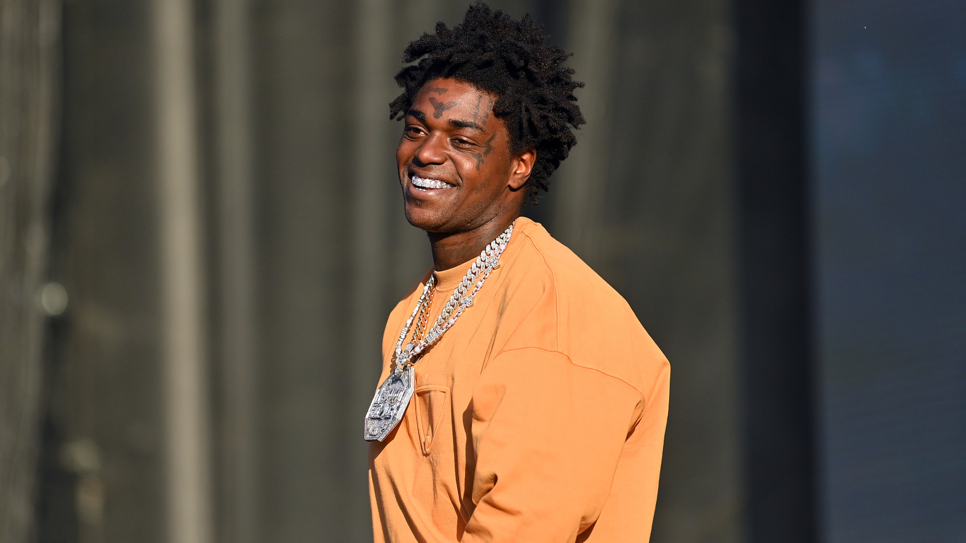 Kodak Black with short dreadlocks wearing an orange shirt and a large chain necklace, smiling outdoors.