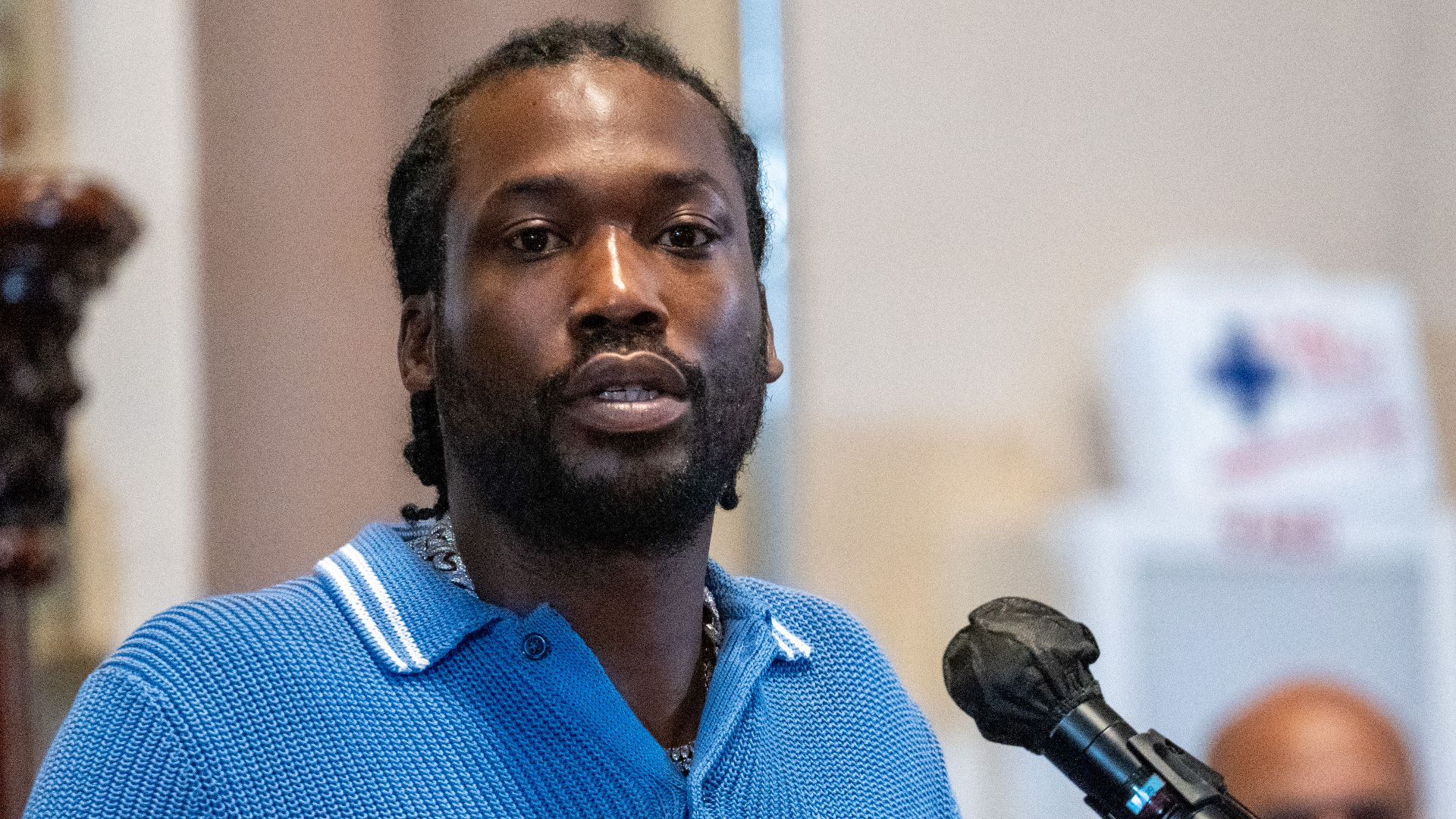 Meek Mill speaks into a microphone, wearing a blue collared shirt in an indoor setting.