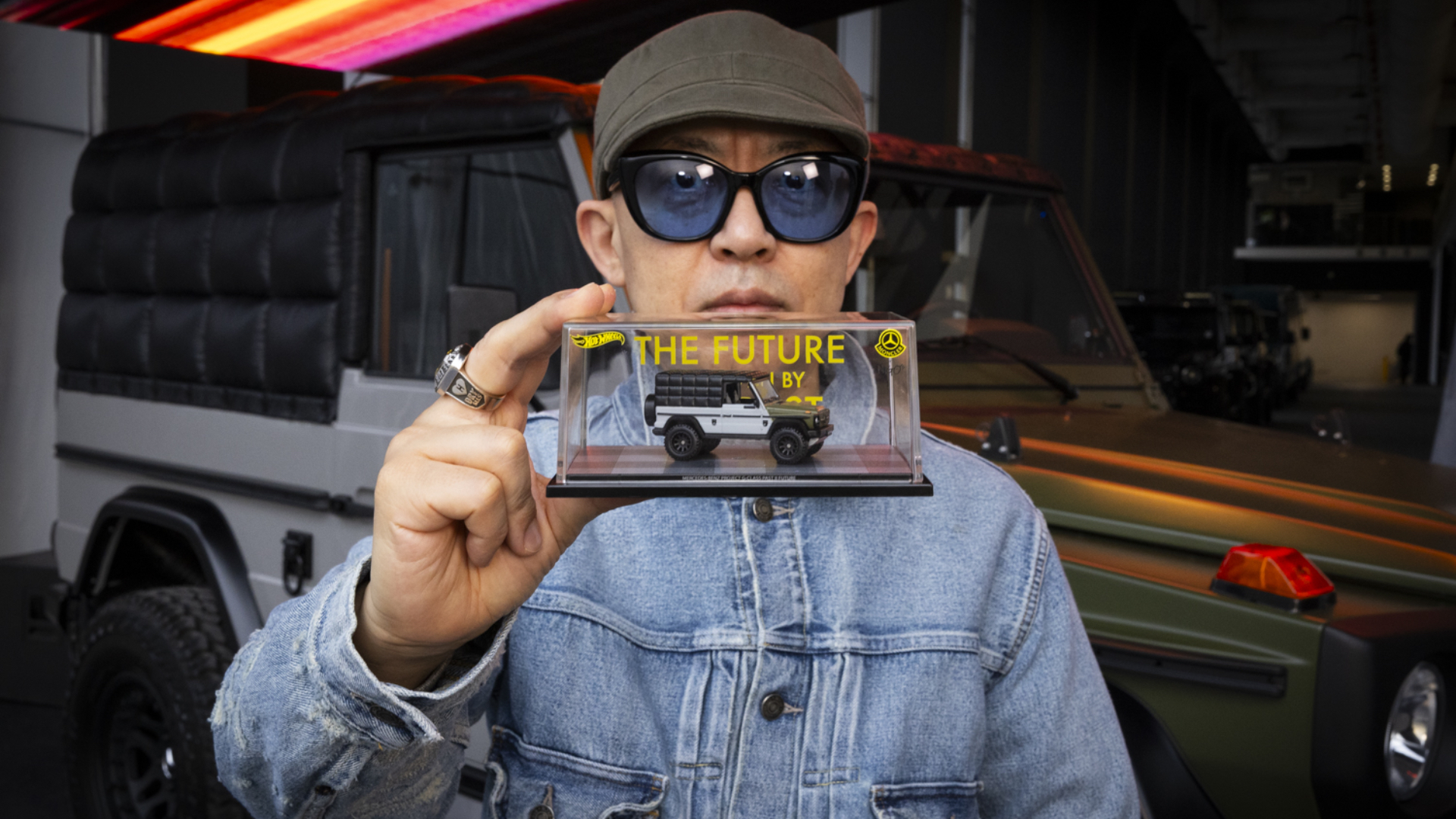 A person wearing sunglasses and a cap holds a model car in a display case labeled "The Future." A vehicle is in the background.