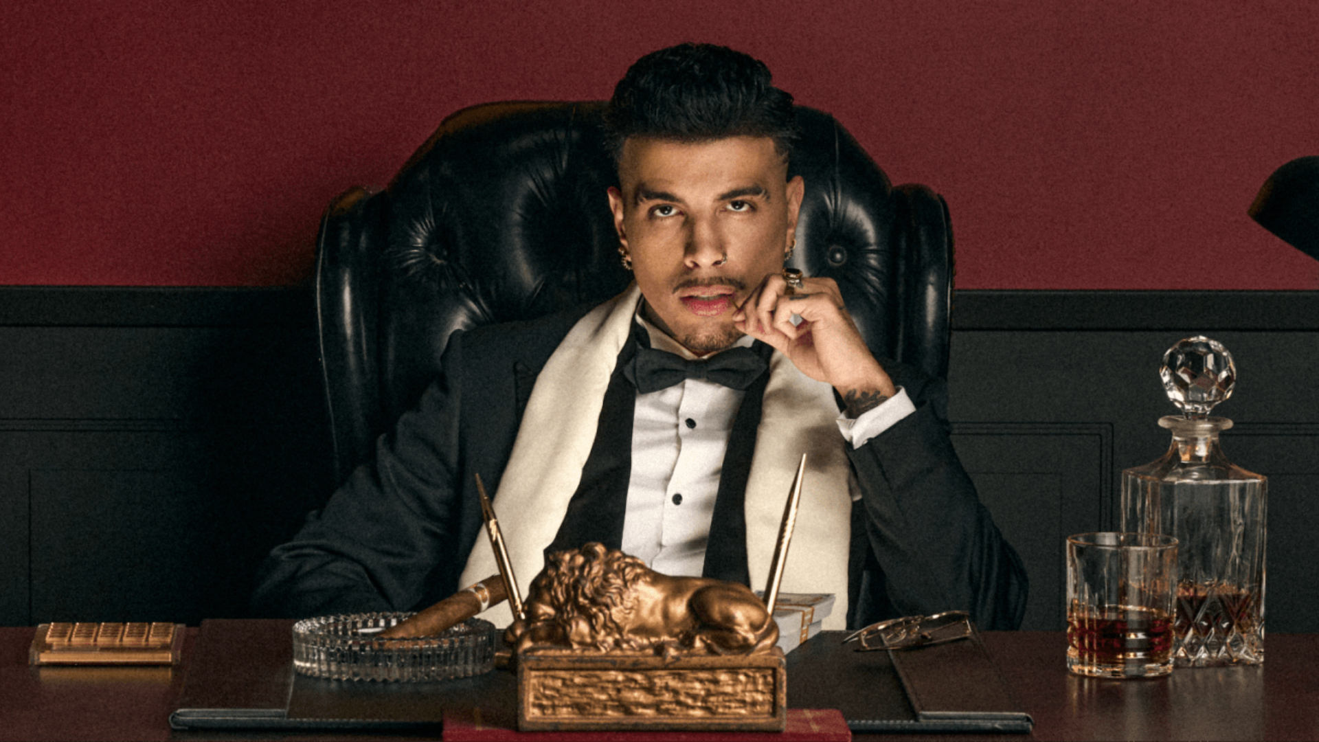 A man in a tuxedo sits at a luxurious desk with a cigar, whiskey, and ornate decor, exuding a sophisticated and contemplative demeanor.