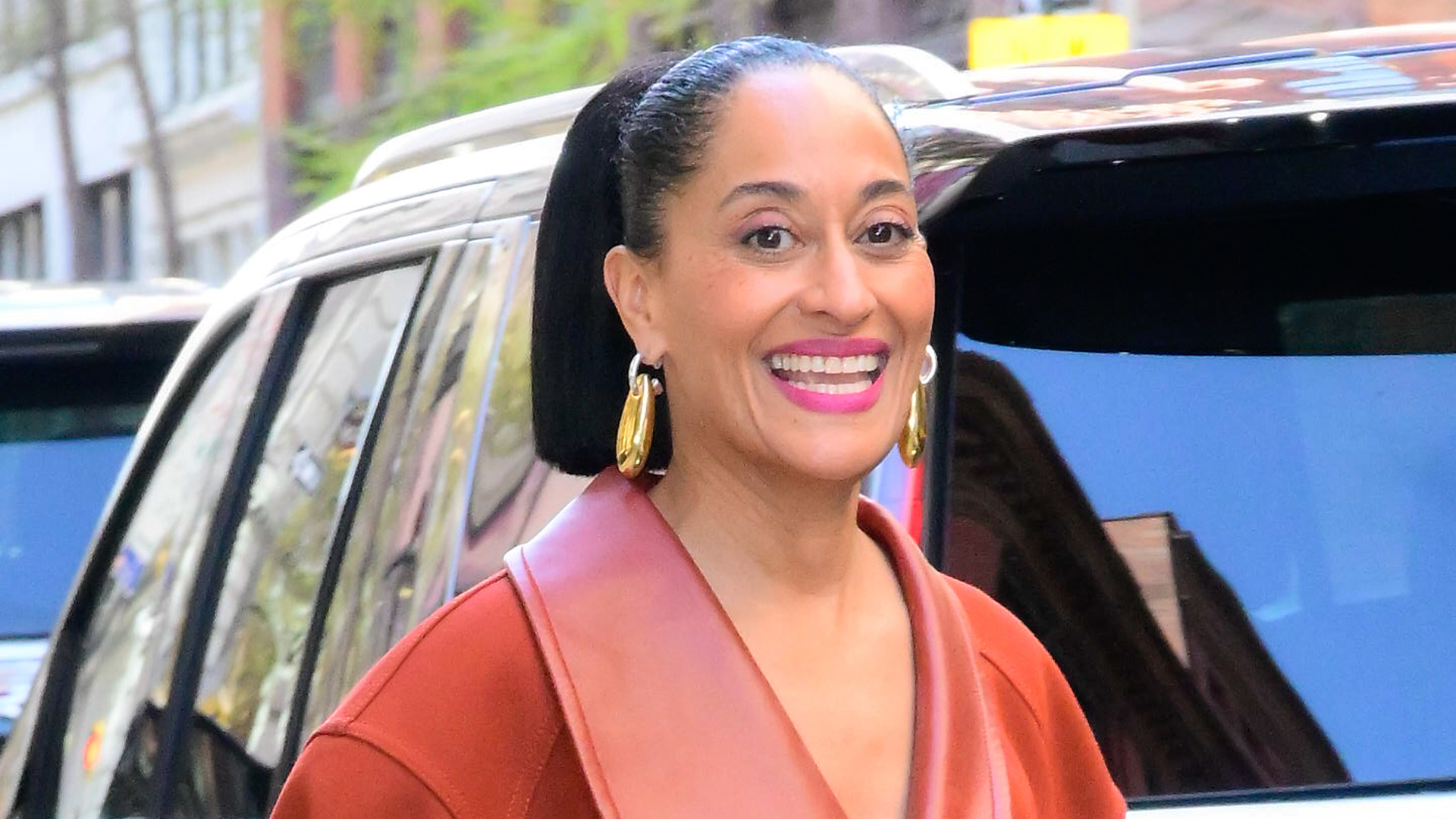 Tracee Ellis Ross smiling, wearing gold hoop earrings and a brown coat, standing near a car on a city street.
