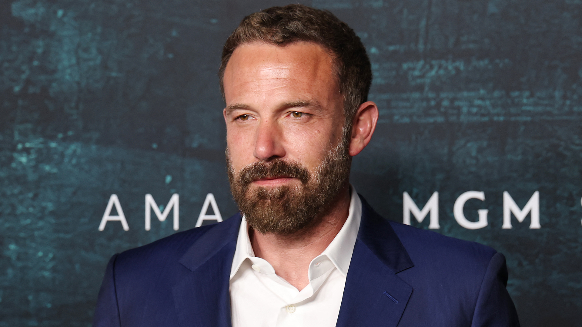 Ben Affleck at an event, wearing a blue suit and white shirt, with a beard and short hair, standing in front of a dark backdrop.