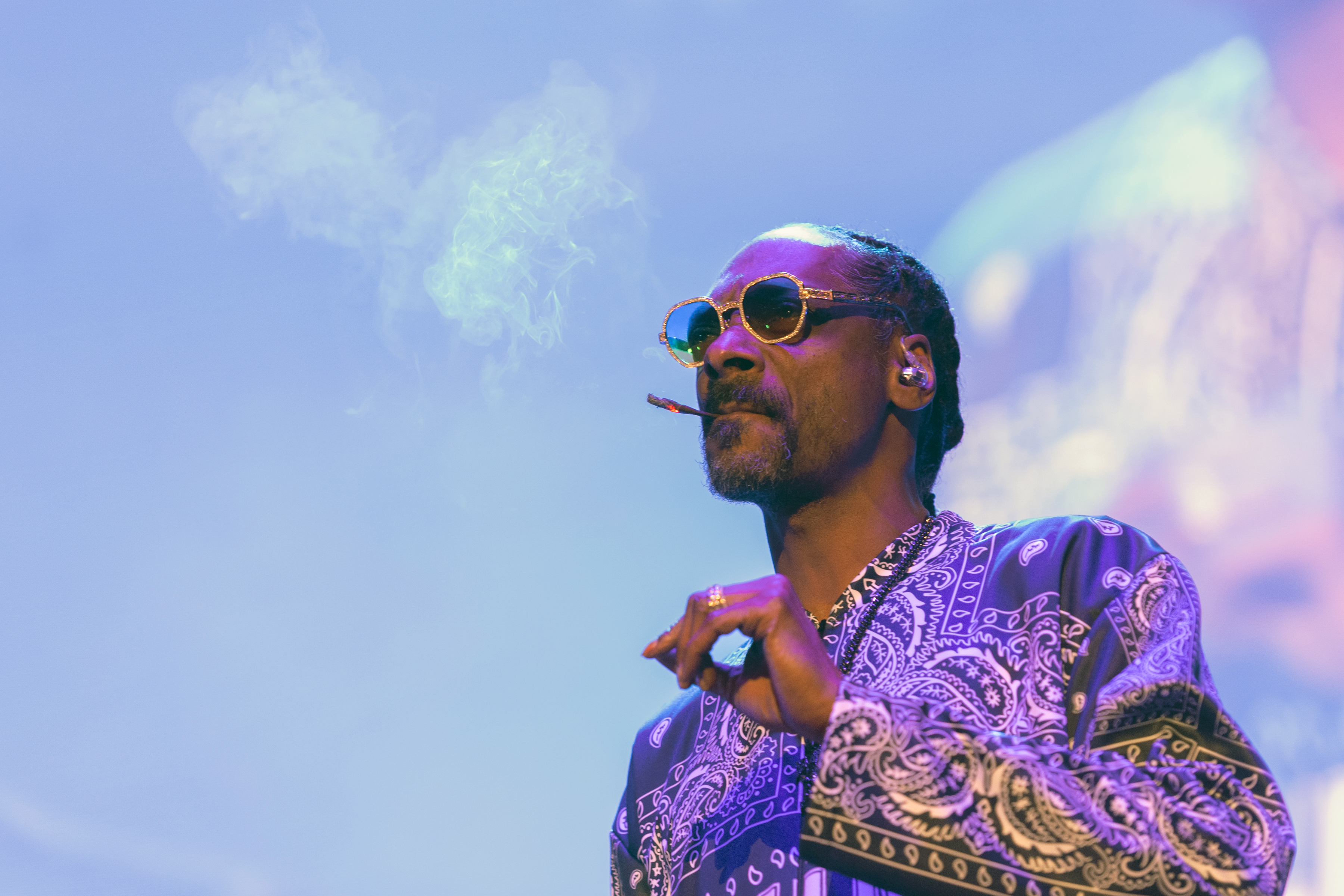Snoop Dogg on stage, wearing sunglasses and a patterned shirt, smoking with a relaxed expression.