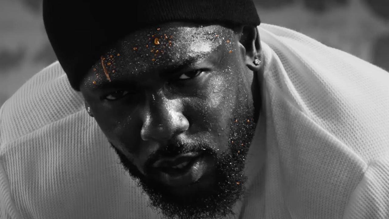 A close-up of Kendrick Lamar wearing a black beanie and white shirt, with gold sweat on his face, looking intently at the camera.