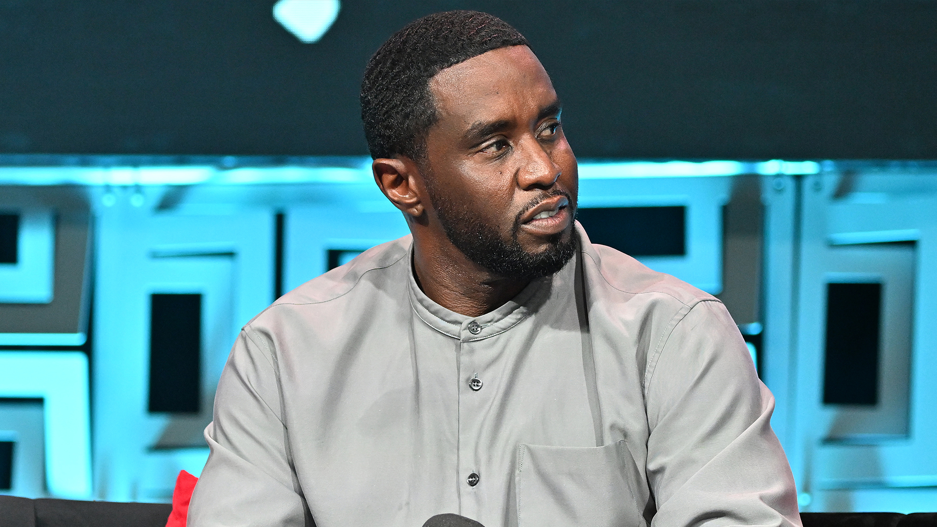 Diddy wearing a gray shirt sits on a stage with a geometric background.