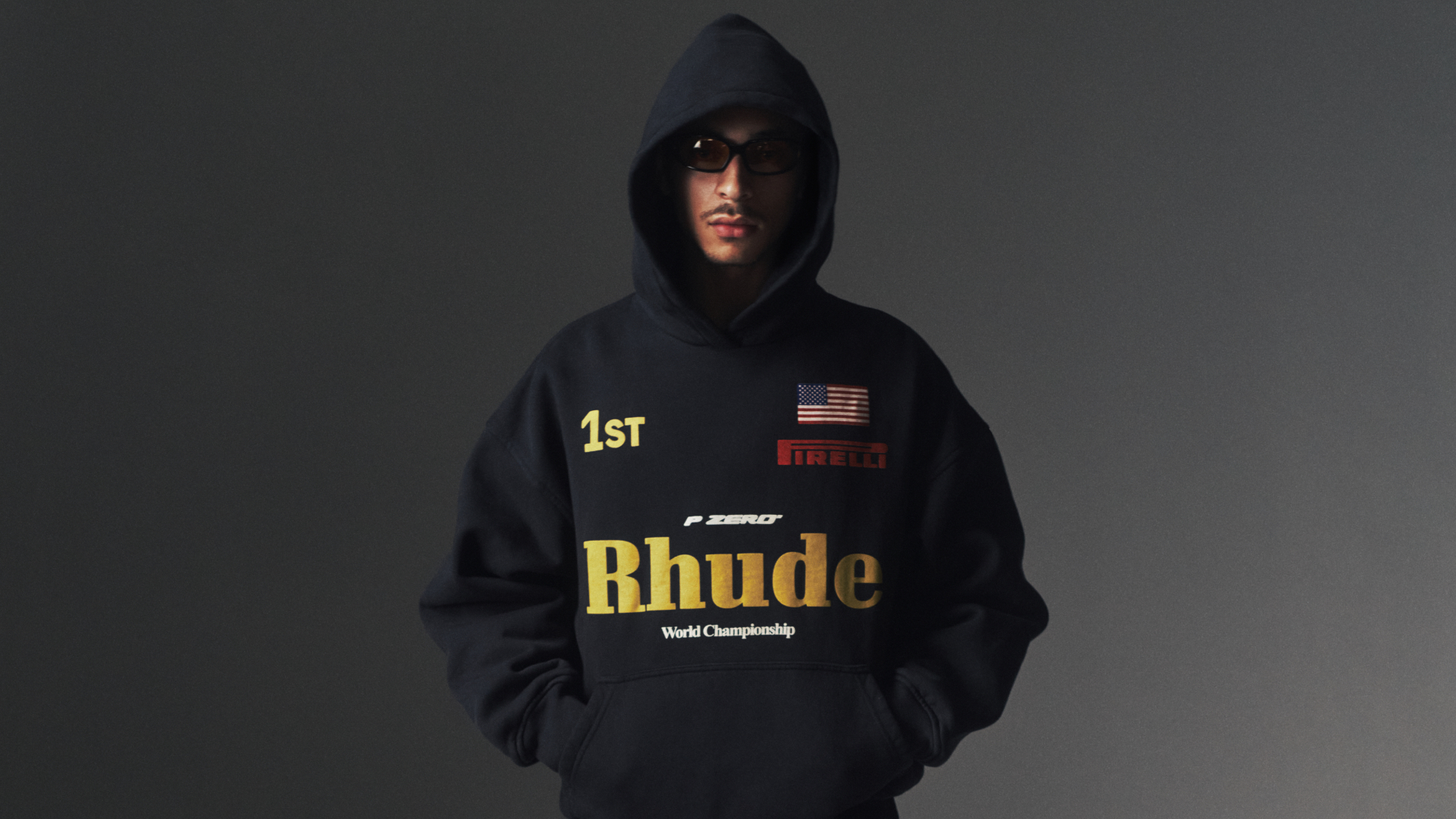 A person wearing a black Rhude hoodie with logos, sunglasses, and a hood, standing against a dark background.