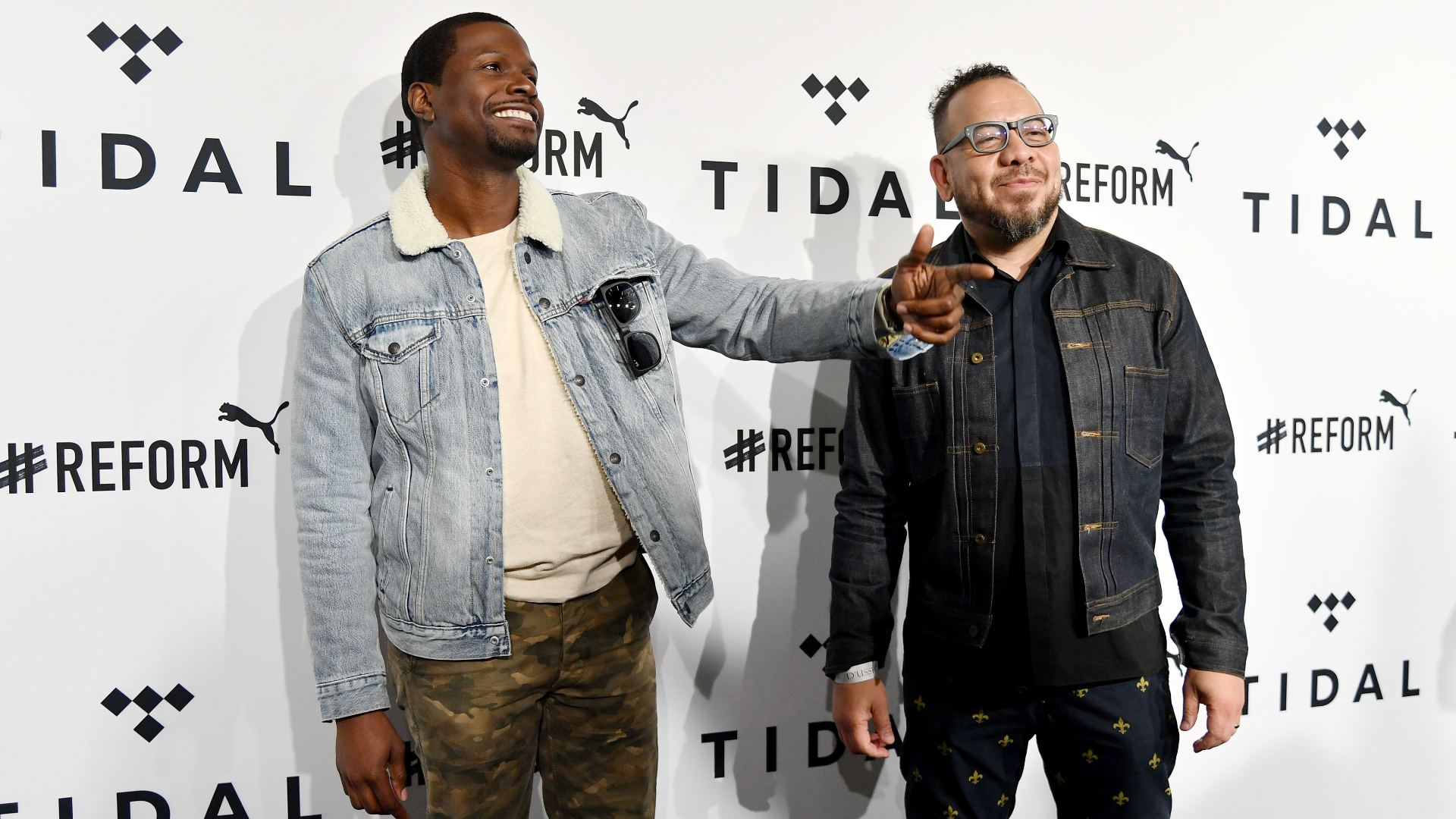 Two men smiling at a TIDAL event, one in a denim jacket and camo pants, the other in a dark jacket with glasses.