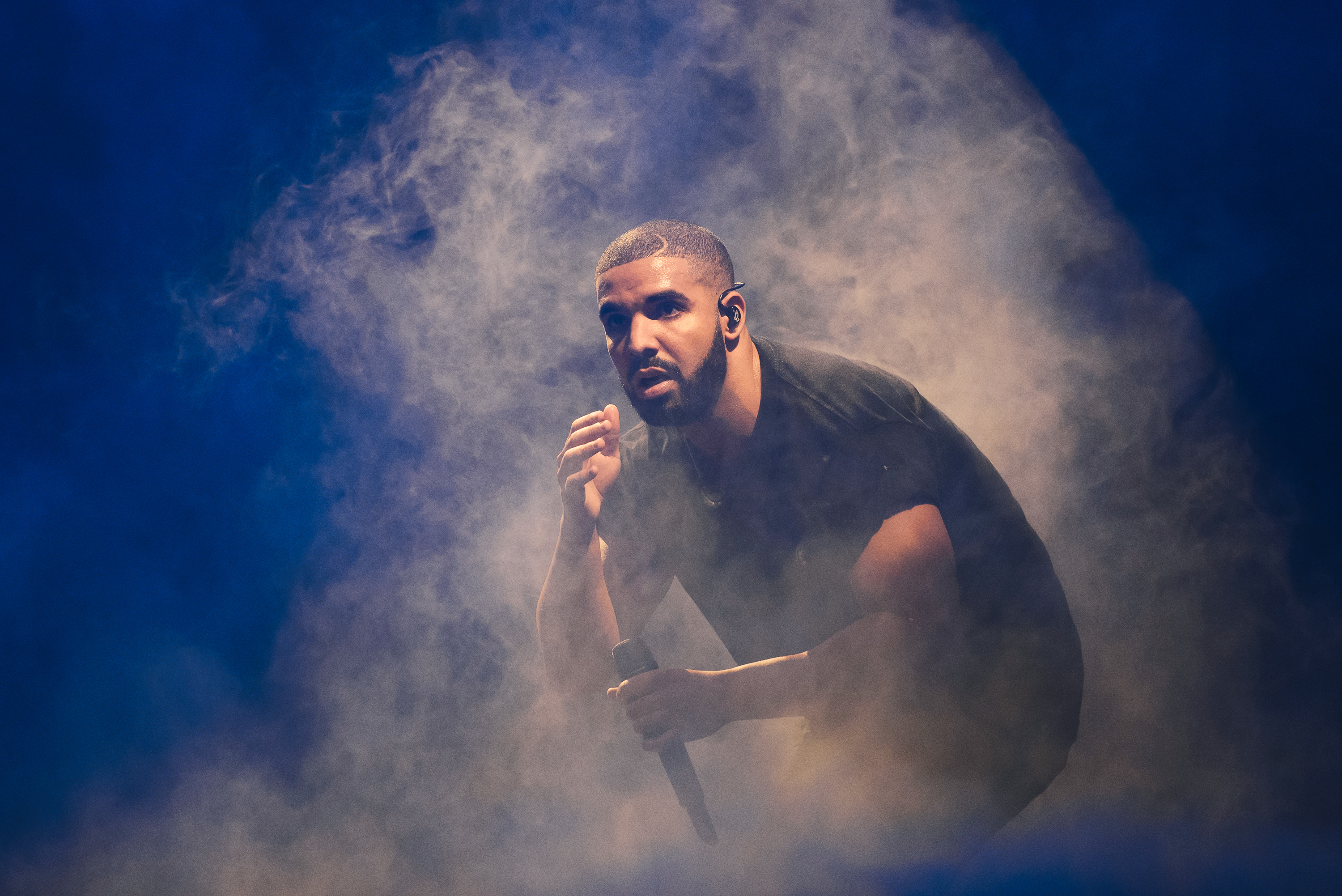 Drake performing on stage, surrounded by smoke, wearing a black shirt and holding a microphone.