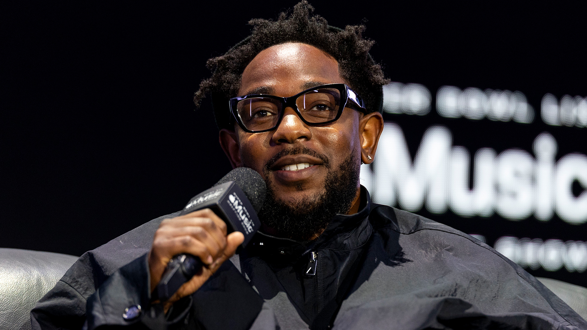 Kendrick Lamar speaking into a microphone, wearing glasses and a dark jacket, seated against a backdrop with blurred text.