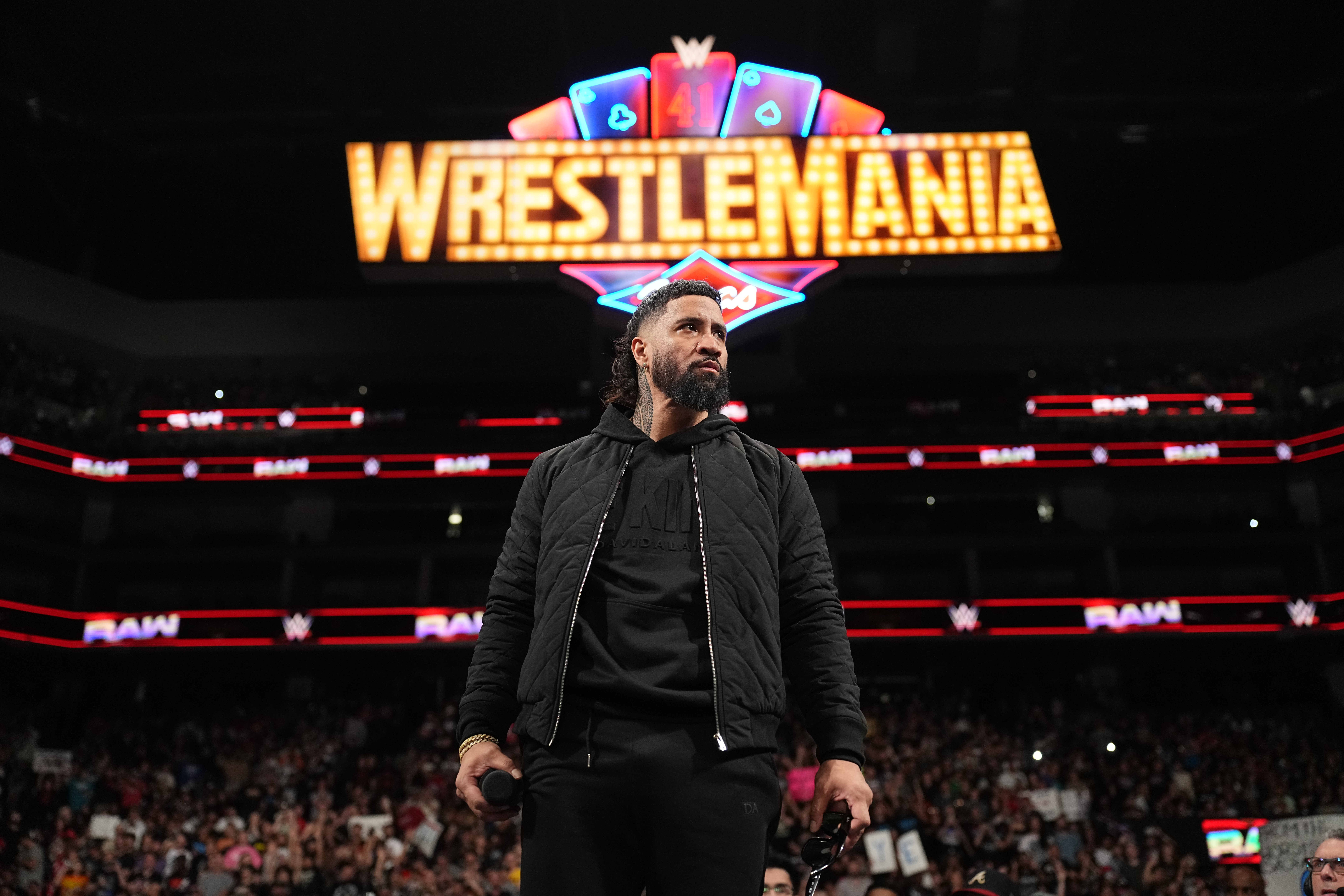 A man with a beard stands in a wrestling arena with a "WrestleMania" sign overhead, holding a microphone, surrounded by a crowd.